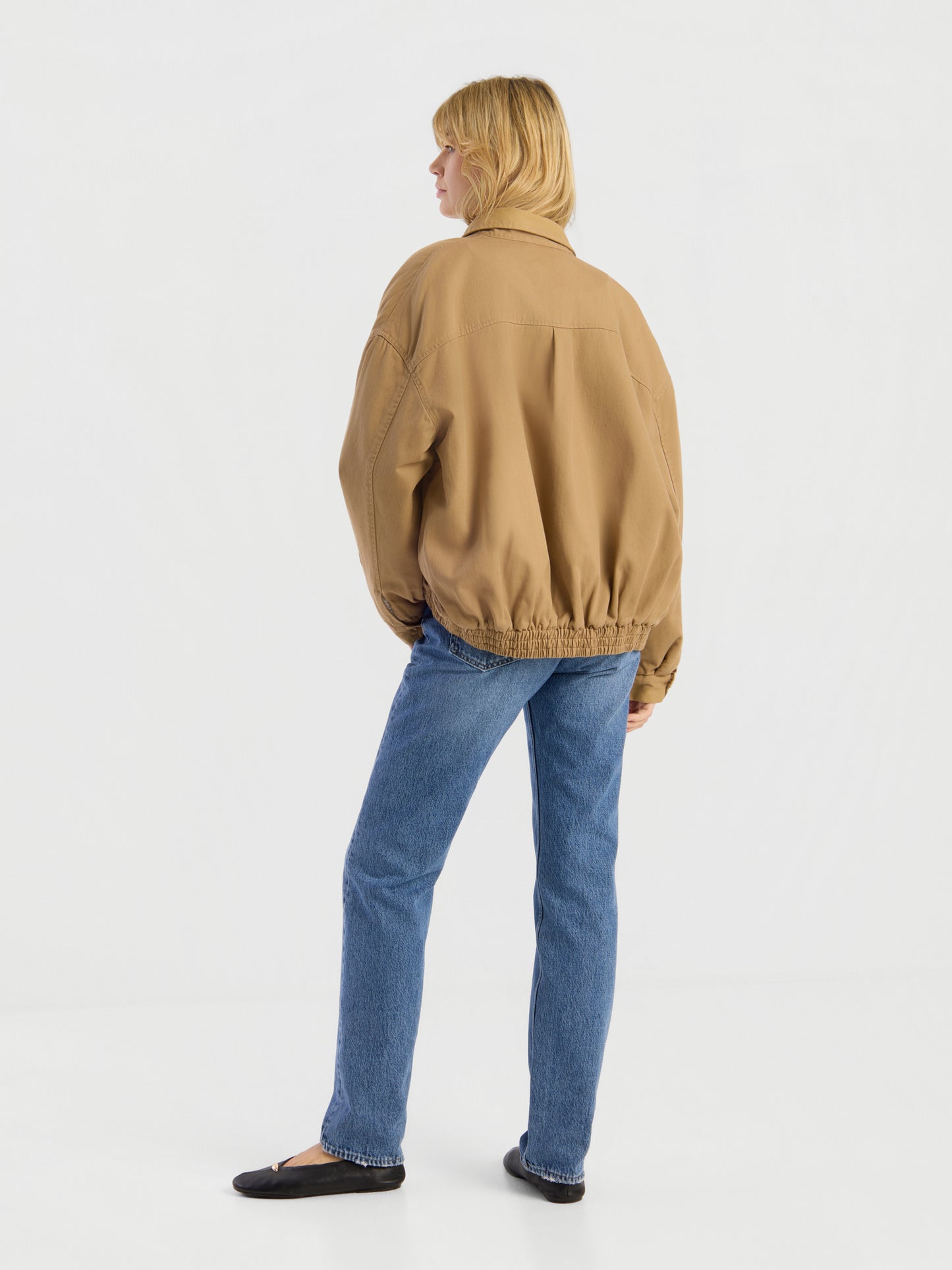 Eine Person mit blonden Haaren, bekleidet mit einer beigen „Button Down Bomber Short“-Jacke, blauen Jeans und schwarzen Schuhen, steht mit dem Rücken zur Kamera vor einem schlichten weißen Hintergrund.