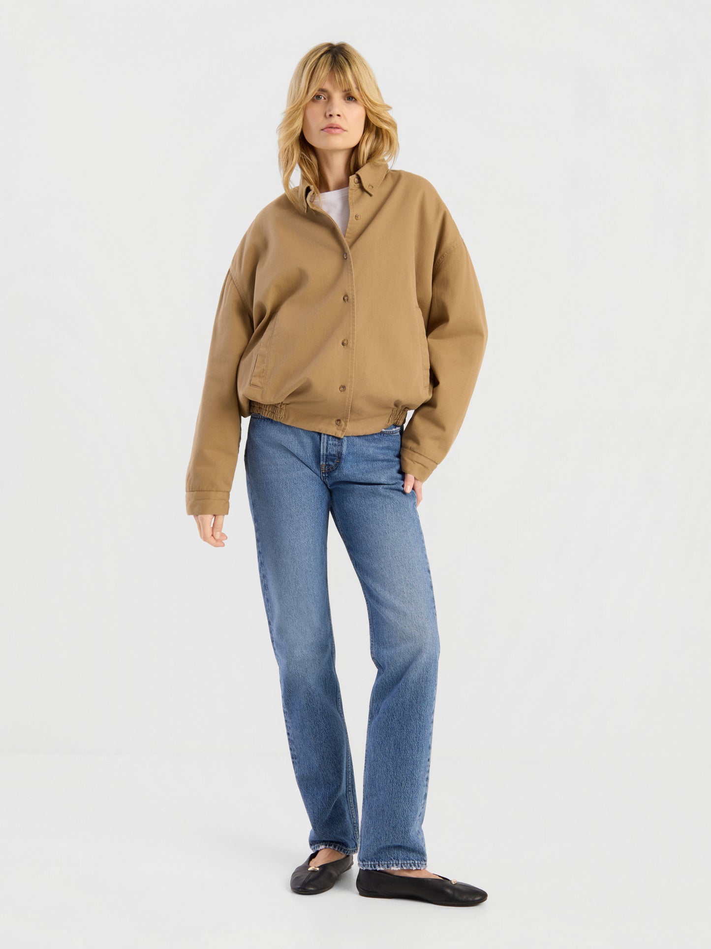 Eine Person mit blonden Haaren trägt die beigefarbene „Button Down Bomber Short“-Jacke, dazu blaue Jeans und schwarze Ballerinas, und steht vor einem schlichten weißen Hintergrund.