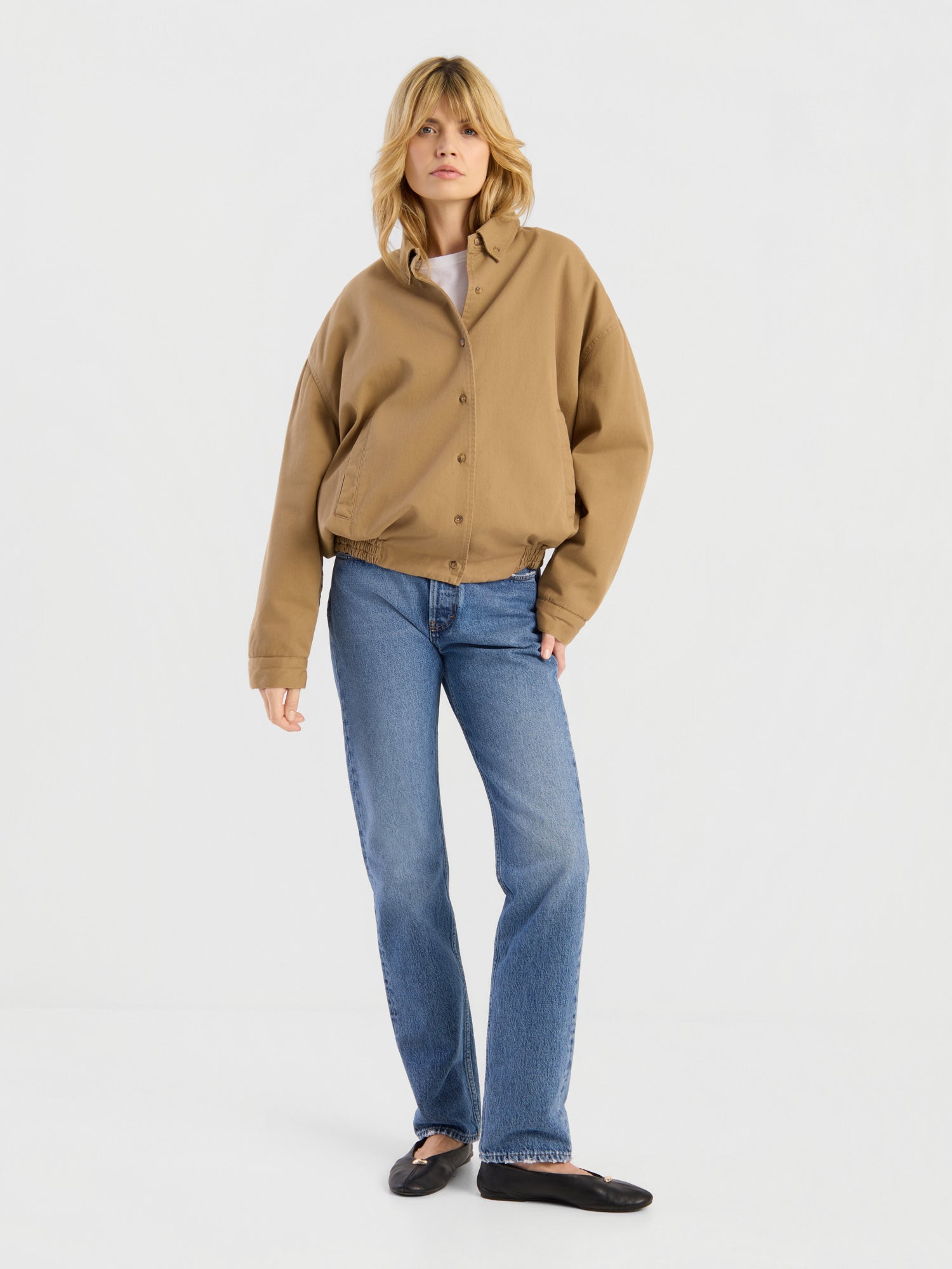 Eine Person mit blonden Haaren trägt die beigefarbene „Button Down Bomber Short“-Jacke, dazu blaue Jeans und schwarze Ballerinas, und steht vor einem schlichten weißen Hintergrund.