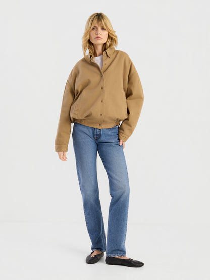 Eine Person mit blonden Haaren trägt die beigefarbene „Button Down Bomber Short“-Jacke, dazu blaue Jeans und schwarze Ballerinas, und steht vor einem schlichten weißen Hintergrund.
