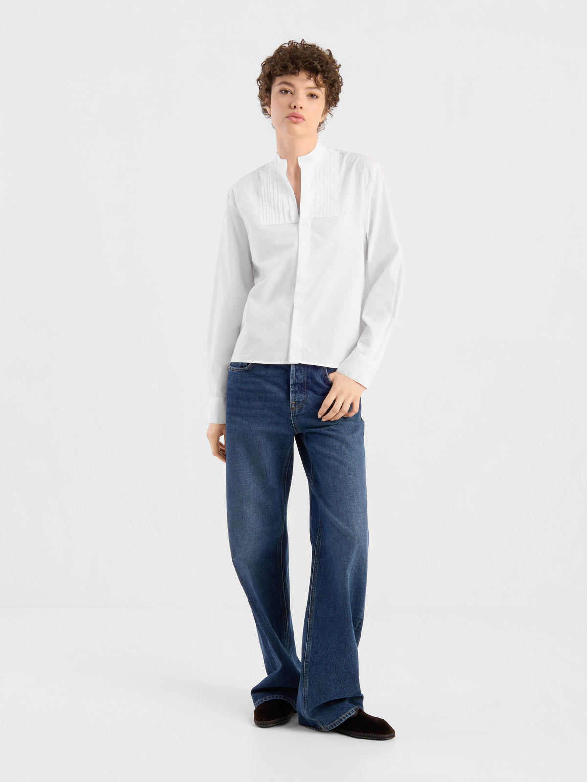 Eine Person mit kurzen Locken präsentiert die Bluse „Tuxedo“ – eine elegante, weiße, langärmelige Baumwollbluse mit Biesen – in Kombination mit einer locker sitzenden blauen Jeans vor einem schlichten weißen Hintergrund. Diese stilvolle Bluse wurde in Portugal meisterhaft gefertigt und sorgt für einen raffinierten Look.