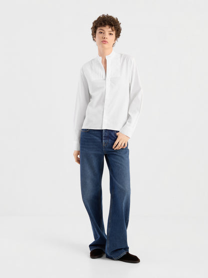 Eine Person mit kurzen Locken präsentiert die Bluse „Tuxedo“ – eine elegante, weiße, langärmelige Baumwollbluse mit Biesen – in Kombination mit einer locker sitzenden blauen Jeans vor einem schlichten weißen Hintergrund. Diese stilvolle Bluse wurde in Portugal meisterhaft gefertigt und sorgt für einen raffinierten Look.