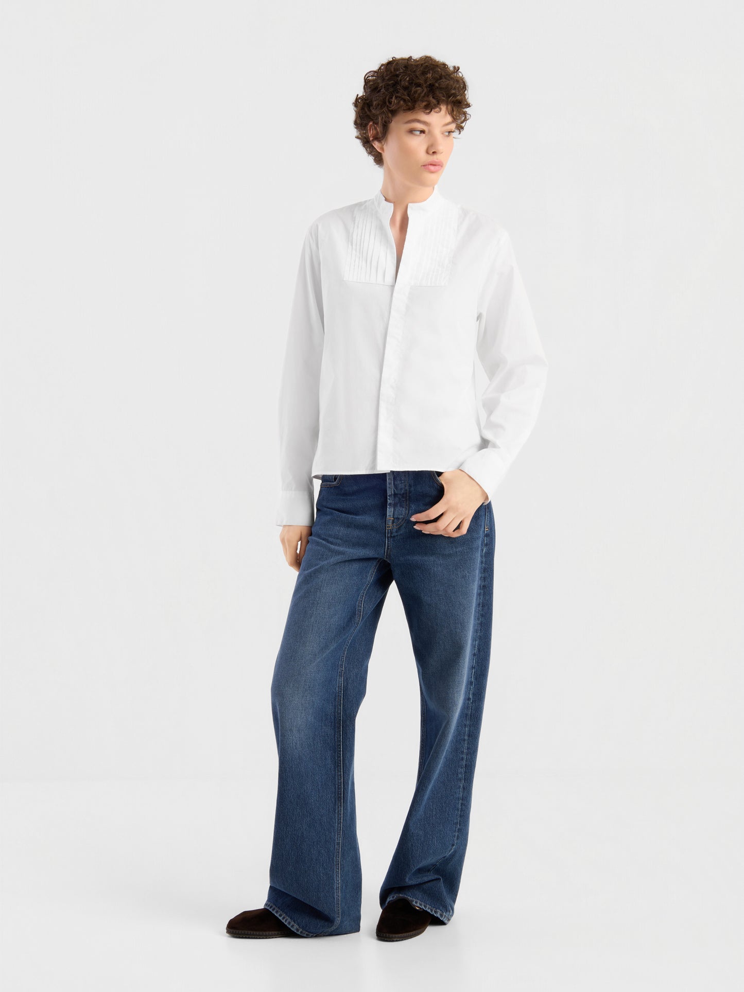 Eine Person, die ein weißes Langarmhemd mit Plissee-Details und die NOLA Jeans „Dark Blue – Wide Leg“ für Damen aus hochwertigem Denim trägt, steht vor einem schlichten weißen Hintergrund, blickt zur Seite und hat eine Hand in der Hosentasche.
