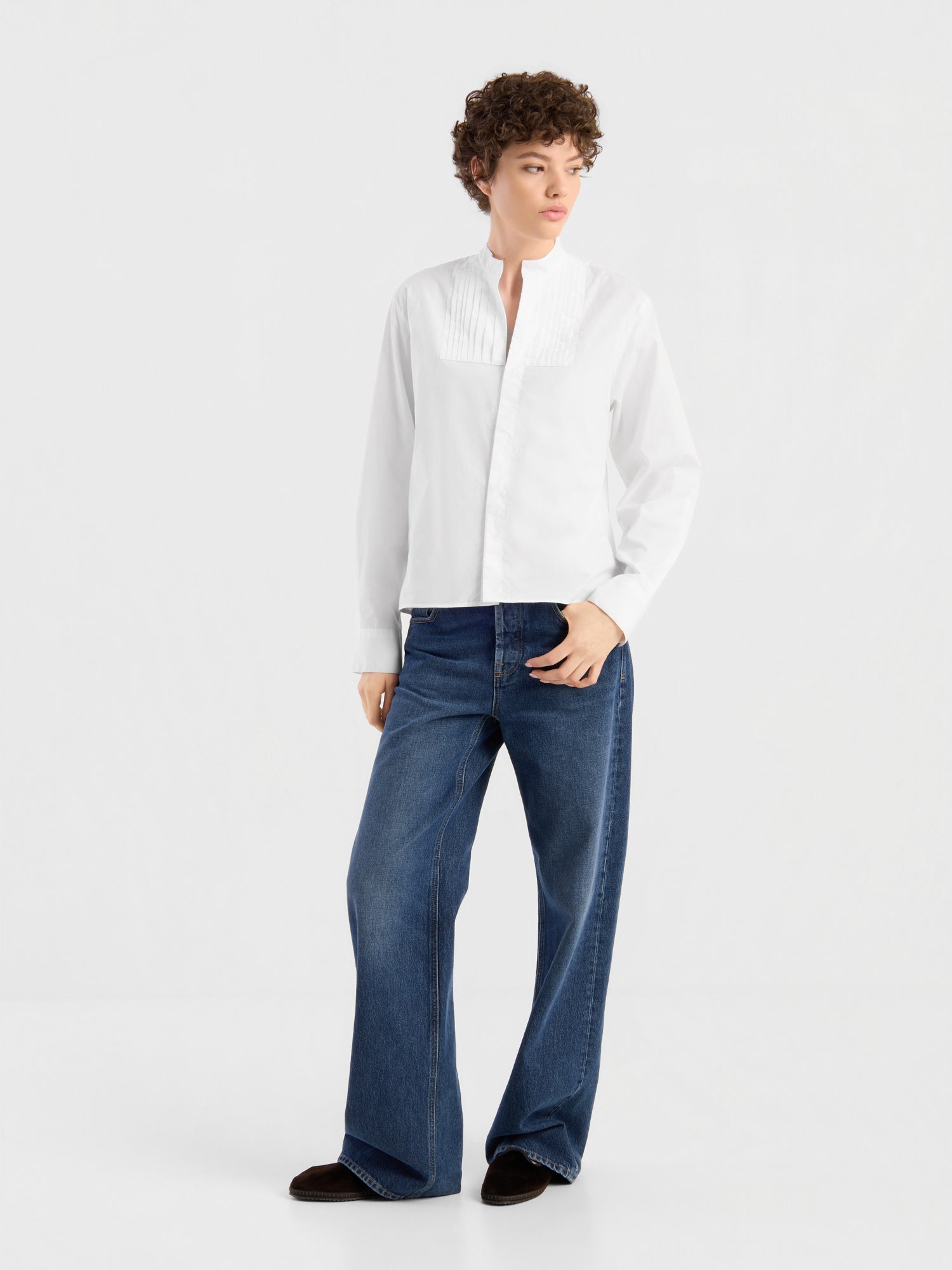 Eine Person, die ein weißes Langarmhemd mit Plissee-Details und die NOLA Jeans „Dark Blue – Wide Leg“ für Damen aus hochwertigem Denim trägt, steht vor einem schlichten weißen Hintergrund, blickt zur Seite und hat eine Hand in der Hosentasche.