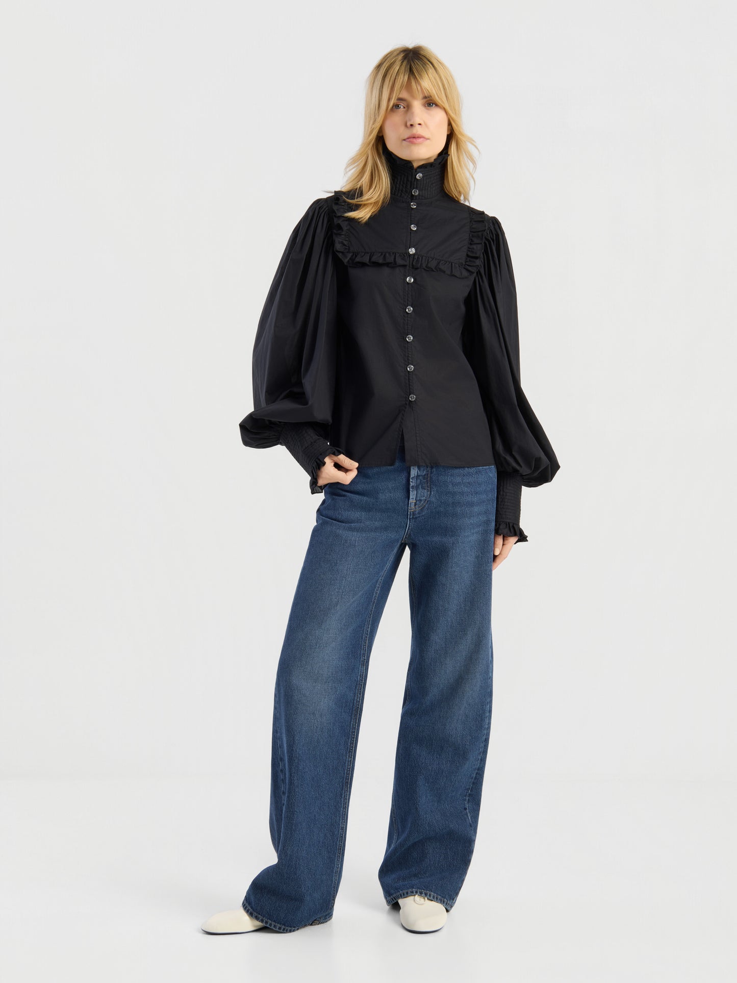Vor einem schlichten weißen Hintergrund steht eine Frau, die die Bluse „Victoria“ trägt, die sich durch Rüschen und voluminöse Ärmel auszeichnet, dazu eine blaue Jeans mit weitem Bein und weiße Schuhe.