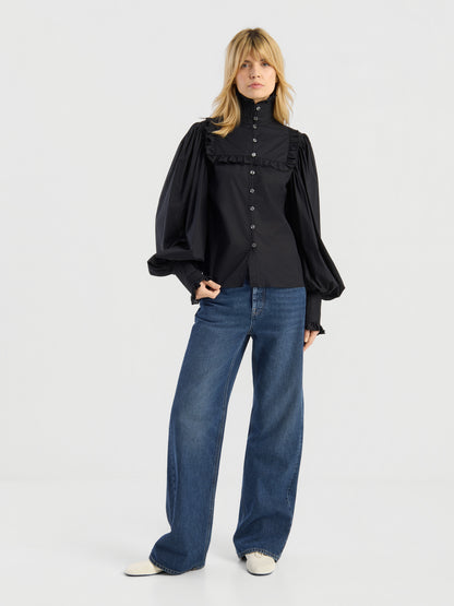 Vor einem schlichten weißen Hintergrund steht eine Frau, die die Bluse „Victoria“ trägt, die sich durch Rüschen und voluminöse Ärmel auszeichnet, dazu eine blaue Jeans mit weitem Bein und weiße Schuhe.