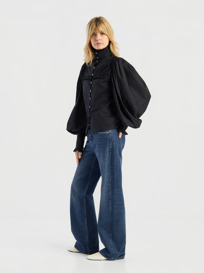 Eine Frau posiert in der „Bluse Victoria“, einer schwarzen Bluse mit voluminösen Ballonärmeln, kombiniert mit einer blauen Wide-Leg-Jeans und weißen Schuhen mit spitzer Zehenpartie. Sie hat schulterlanges blondes Haar und steht vor einem schlichten, hellen Hintergrund.