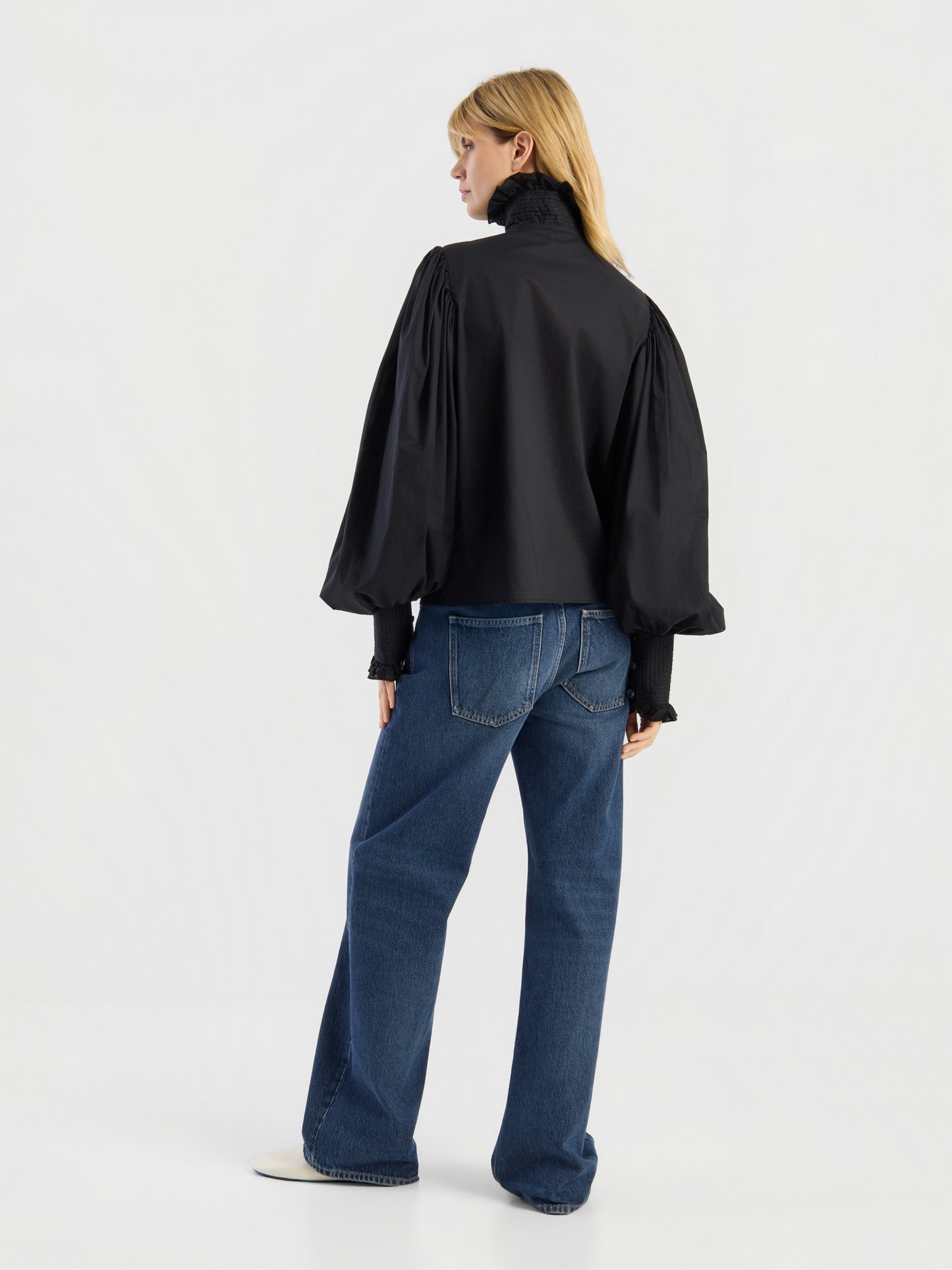 Vor einem schlichten weißen Hintergrund steht eine blonde Person mit dem Rücken zur Kamera und trägt die „Bluse Victoria“ – eine schwarze Bluse mit hohem Kragen und Puffärmeln –, dazu eine blaue Jeans mit weitem Bein und weiße Schuhe.