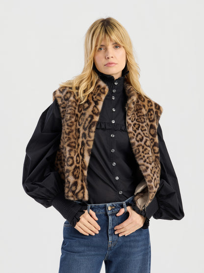 Eine Frau mit blonden Haaren trägt die „Weste Leopard“ – eine Kunstfellweste mit auffälligem Leopardenmuster – über einer schwarzen Bluse mit Knöpfen und Puffärmeln sowie einer blauen Jeans und steht vor einem schlichten, hellen Hintergrund.