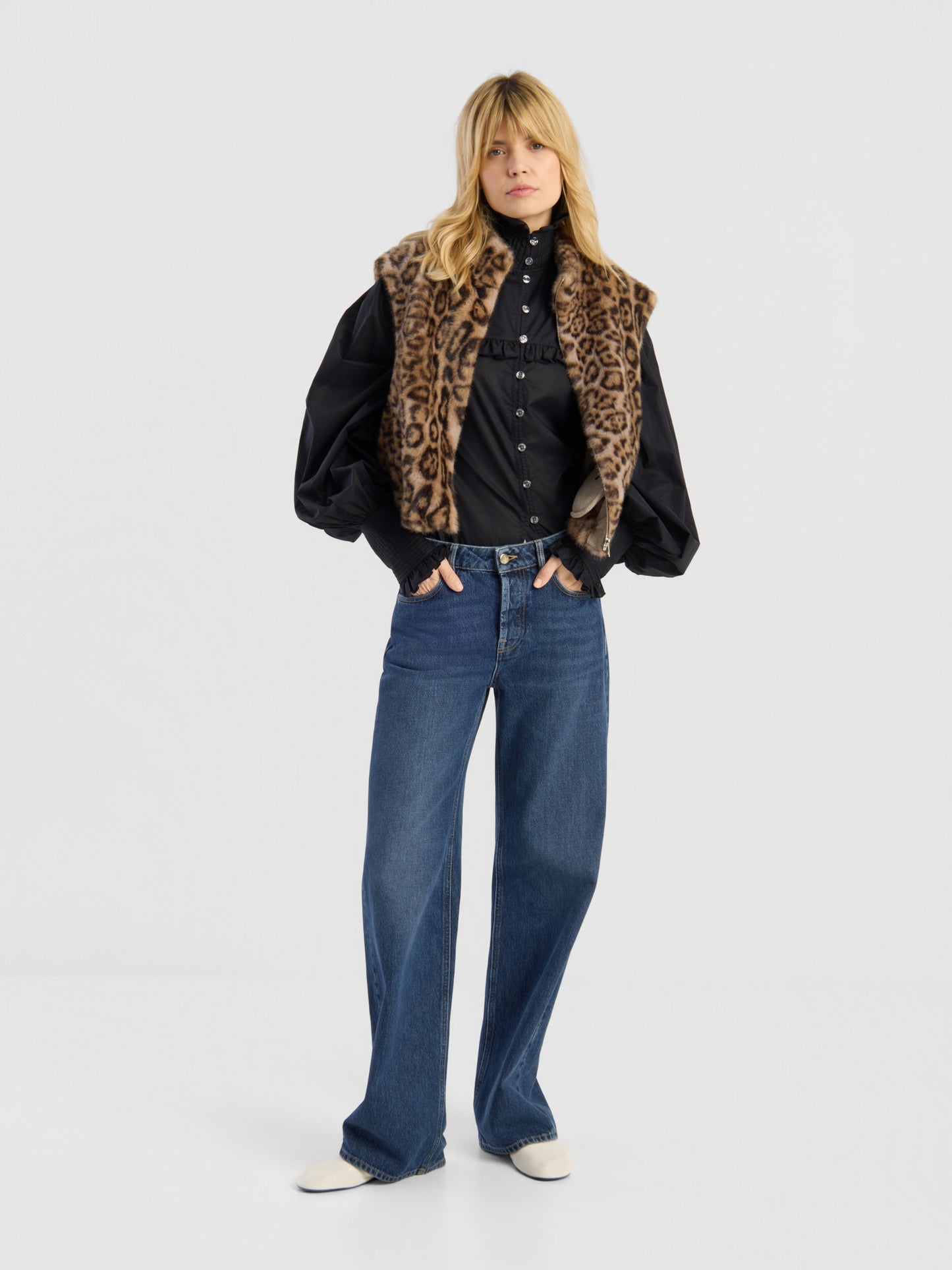 Eine Person steht vor einem schlichten Hintergrund und trägt die „Weste Leopard“ über einem schwarzen Hemd mit Knöpfen und Puffärmeln, einer blauen Jeans mit weitem Bein und weißen Schuhen.