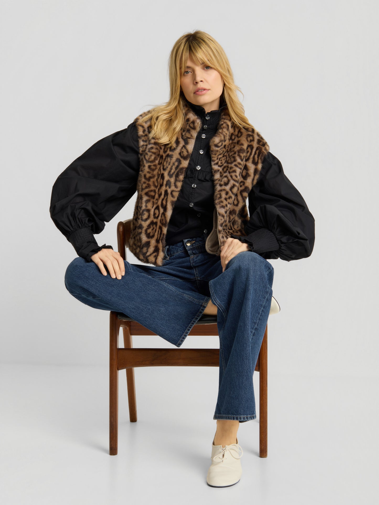 Eine Frau sitzt auf einem Holzstuhl und trägt die „Weste Leopard“, eine schwarze Bluse mit Puffärmeln, eine blaue Jeans und weiße Schuhe – ein Bein hat sie über den Stuhl geschlagen, der andere Fuß ruht auf dem Boden.
