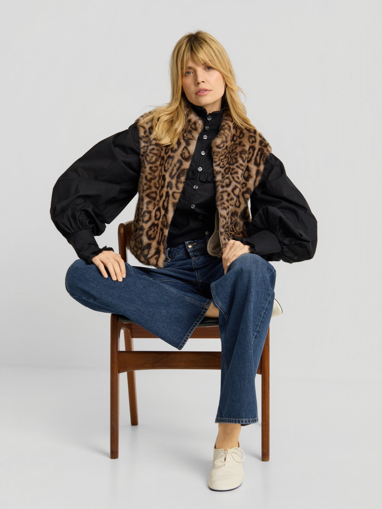 Eine Frau sitzt auf einem Holzstuhl und trägt die „Weste Leopard“, eine schwarze Bluse mit Puffärmeln, eine blaue Jeans und weiße Schuhe – ein Bein hat sie über den Stuhl geschlagen, der andere Fuß ruht auf dem Boden.
