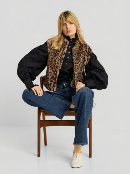 Eine Frau sitzt auf einem Holzstuhl und trägt die „Weste Leopard“, eine schwarze Bluse mit Puffärmeln, eine blaue Jeans und weiße Schuhe – ein Bein hat sie über den Stuhl geschlagen, der andere Fuß ruht auf dem Boden.
