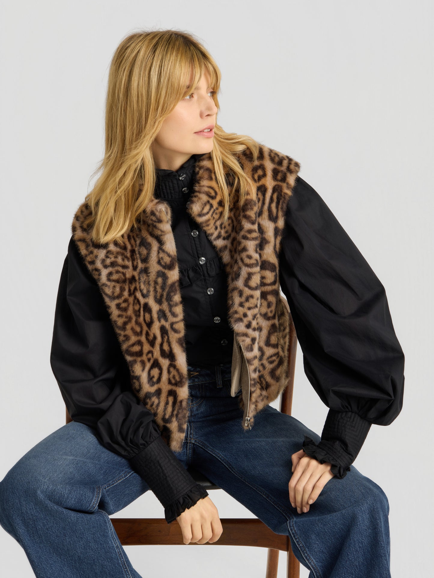 Eine Frau mit blonden Haaren sitzt vor einem schlichten Hintergrund und blickt nach links. Sie trägt die „Weste Leopard“ über einer schwarzen Bluse mit Puffärmeln und einer blau-weit geschnittenen Jeans.