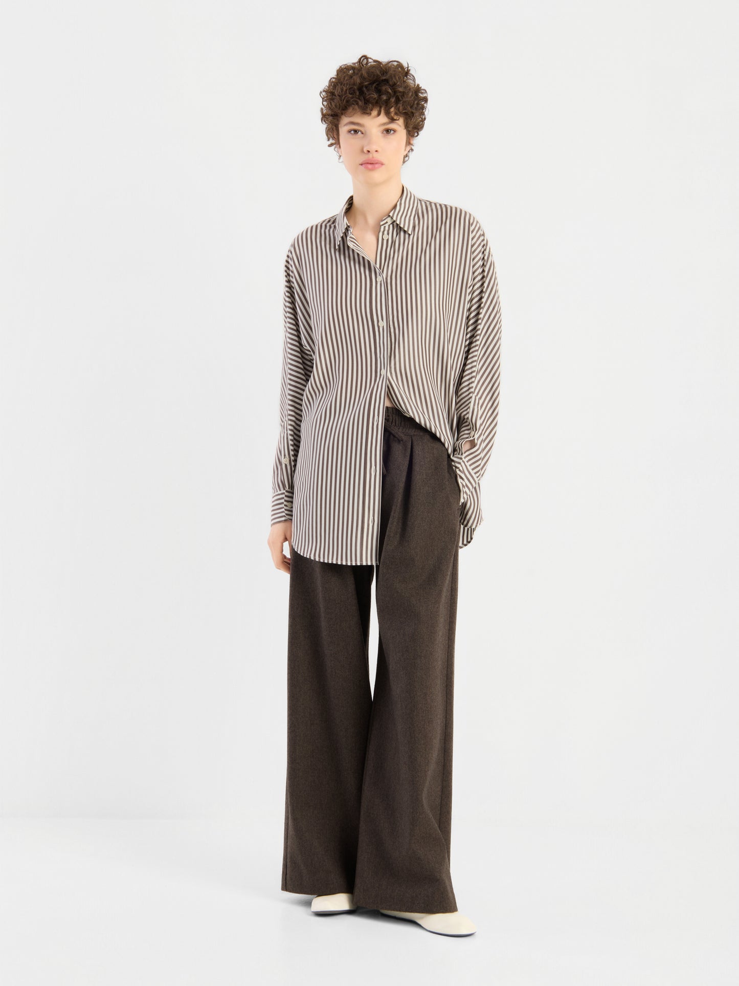 Eine Person mit kurzen Locken trägt das „Bluse Husband Big Stripes“ – ein übergroßes, beige-braun vertikal gestreiftes Hemd mit Knöpfen und breiten Streifen –, dazu eine lockere, dunkelbraune Hose mit weitem Bein und weiße Schuhe; sie steht vor einem schlichten weißen Hintergrund.