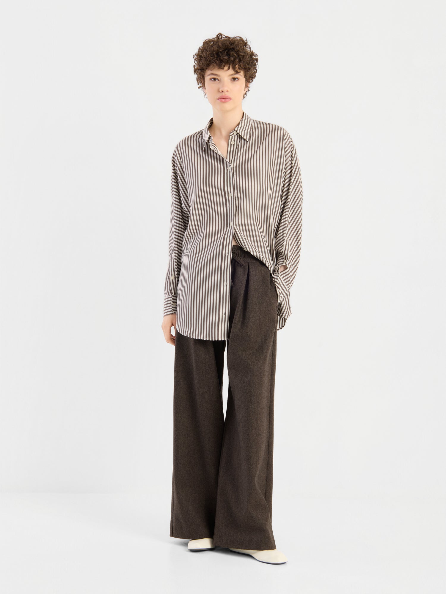 Eine Person mit kurzen Locken trägt das „Bluse Husband Big Stripes“ – ein übergroßes, beige-braun vertikal gestreiftes Hemd mit Knöpfen und breiten Streifen –, dazu eine lockere, dunkelbraune Hose mit weitem Bein und weiße Schuhe; sie steht vor einem schlichten weißen Hintergrund.