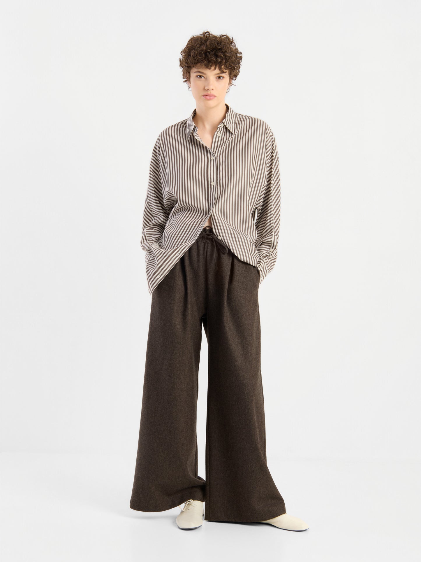 Eine Person mit kurzen Locken, die eine locker sitzende Bluse „Husband Big Stripes“ in Oversize-Passform, eine dunkelbraune Hose mit weitem Bein und cremefarbene Schuhe trägt, steht mit den Händen in den Taschen vor einem schlichten weißen Hintergrund.