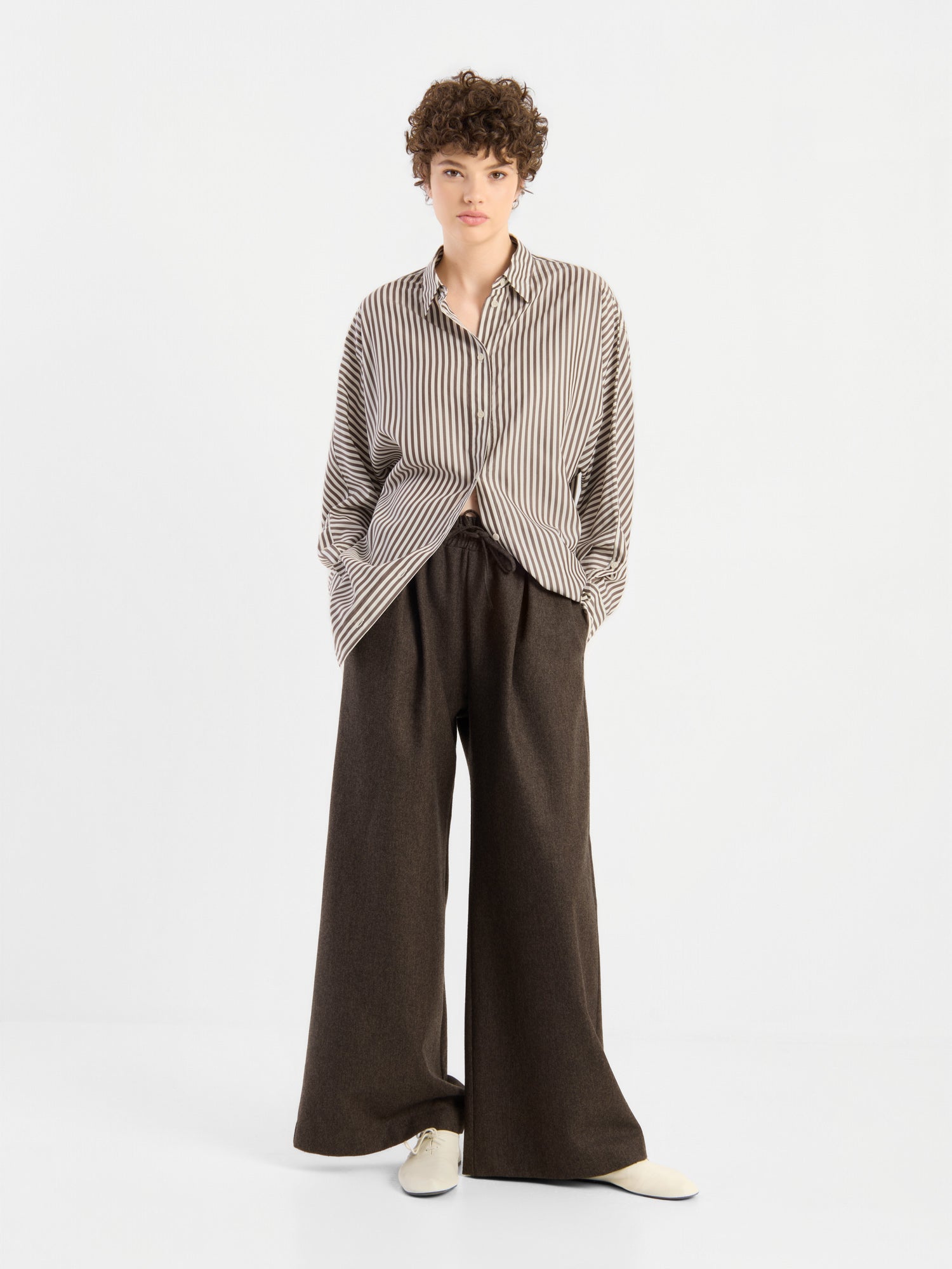 Eine Person mit kurzen Locken, die eine locker sitzende Bluse „Husband Big Stripes“ in Oversize-Passform, eine dunkelbraune Hose mit weitem Bein und cremefarbene Schuhe trägt, steht mit den Händen in den Taschen vor einem schlichten weißen Hintergrund.