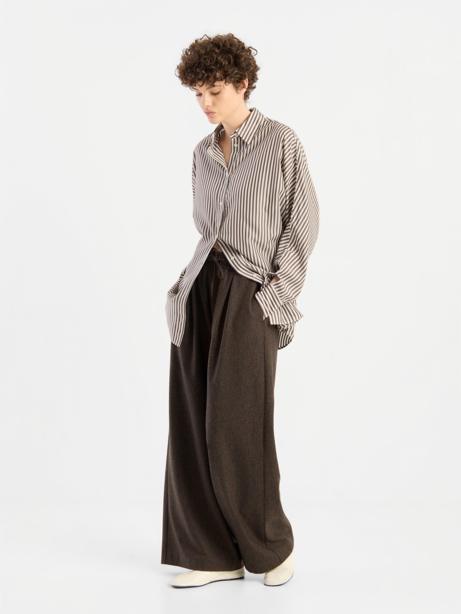 Eine Person, die das „Bluse Husband Big Stripes“ trägt – ein Oversize-Hemd mit breiten Streifen –, kombiniert mit einer braunen Hose mit weitem Bein und weißen Schuhen; sie steht vor einem schlichten weißen Hintergrund, blickt nach unten und hat die Hände in den Taschen.