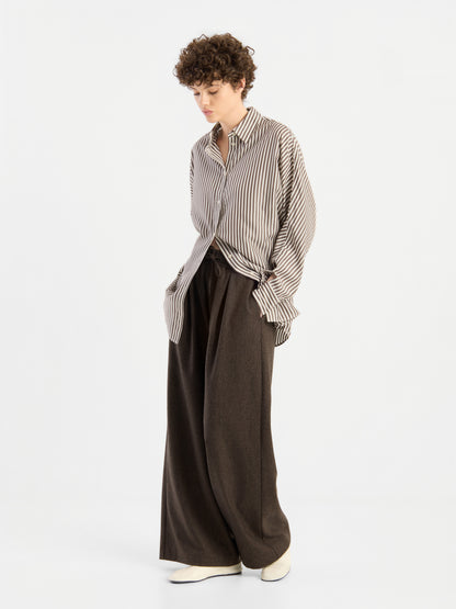 Eine Person, die das „Bluse Husband Big Stripes“ trägt – ein Oversize-Hemd mit breiten Streifen –, kombiniert mit einer braunen Hose mit weitem Bein und weißen Schuhen; sie steht vor einem schlichten weißen Hintergrund, blickt nach unten und hat die Hände in den Taschen.