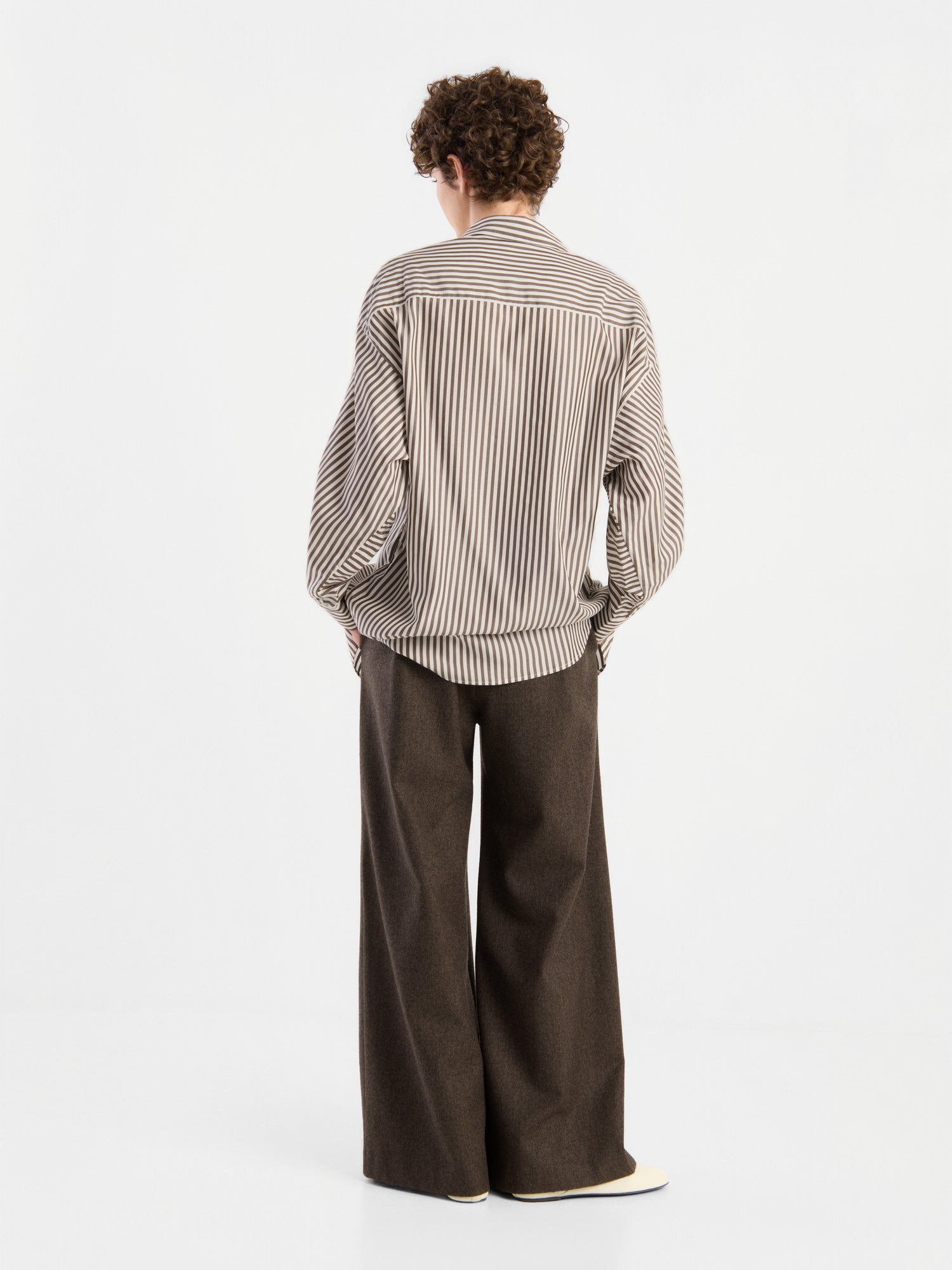 Eine Person steht mit dem Rücken zur Kamera und trägt eine Bluse „Husband Big Stripes“ in Oversize-Passform sowie eine braune Hose mit weitem Bein, vor einem schlichten weißen Hintergrund.