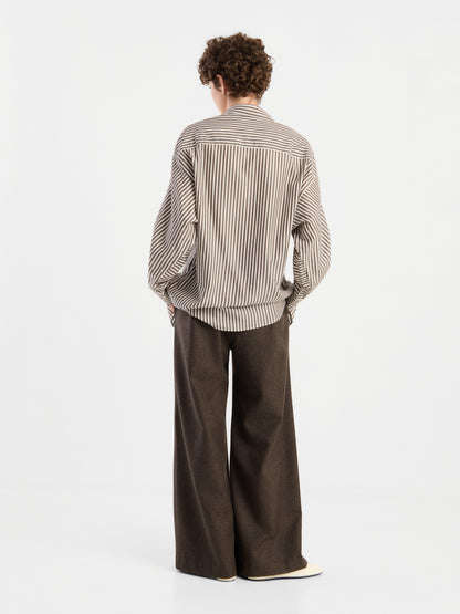 Eine Person steht mit dem Rücken zur Kamera und trägt eine Bluse „Husband Big Stripes“ in Oversize-Passform sowie eine braune Hose mit weitem Bein, vor einem schlichten weißen Hintergrund.