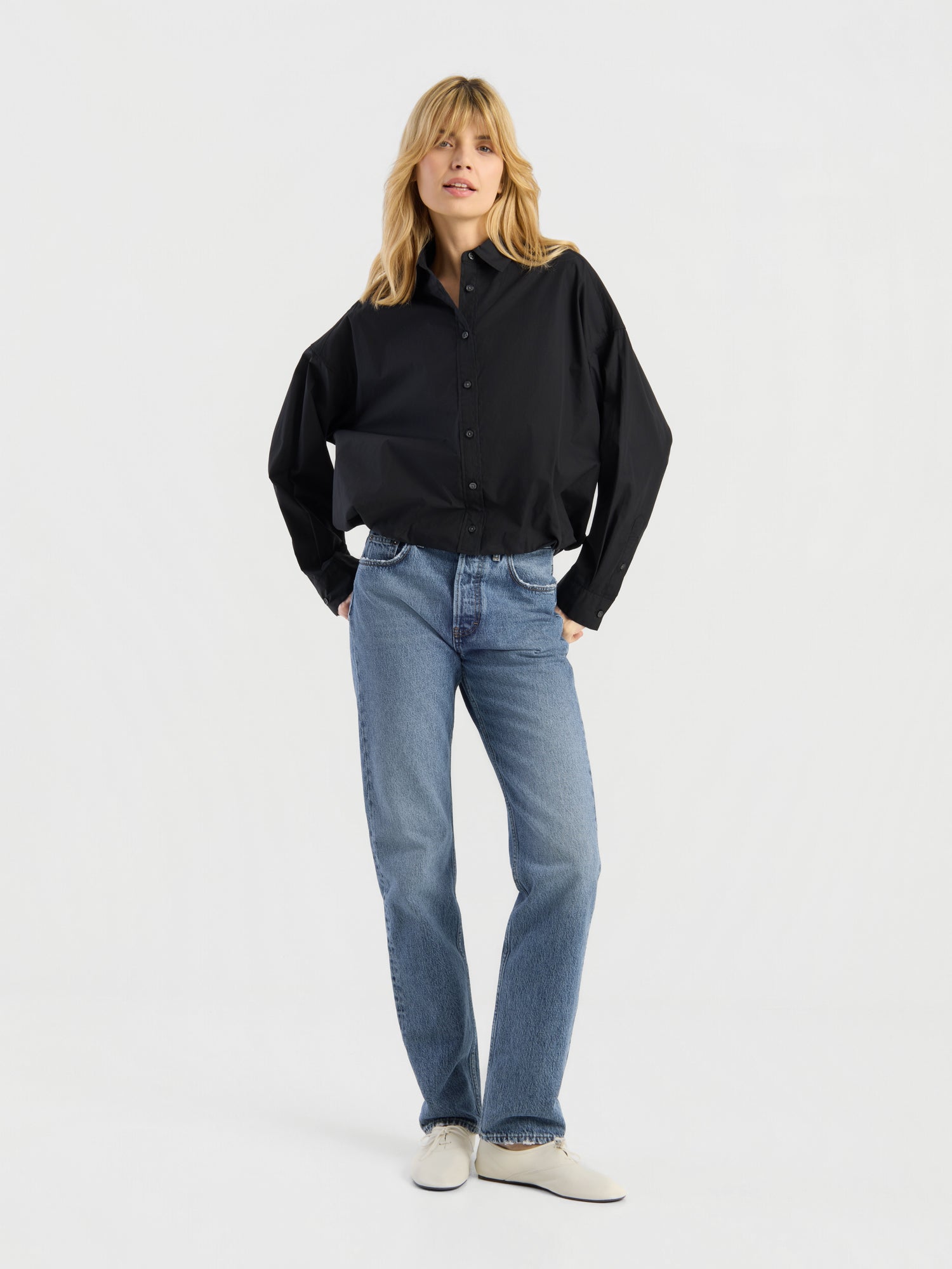 Eine Person mit blonden Haaren, gekleidet in ein locker sitzendes schwarzes Hemd mit Knöpfen, eine „SIENNA Jeans Light Blue – Straight Leg Damenjeans“ aus hochwertigem Denim sowie weiße Schuhe, steht vor einem schlichten weißen Hintergrund, die Hände in den Taschen, und präsentiert dabei mühelos diese stilvolle Damenjeans.