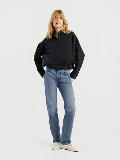 Eine Person mit blonden Haaren, gekleidet in ein locker sitzendes schwarzes Hemd mit Knöpfen, eine „SIENNA Jeans Light Blue – Straight Leg Damenjeans“ aus hochwertigem Denim sowie weiße Schuhe, steht vor einem schlichten weißen Hintergrund, die Hände in den Taschen, und präsentiert dabei mühelos diese stilvolle Damenjeans.