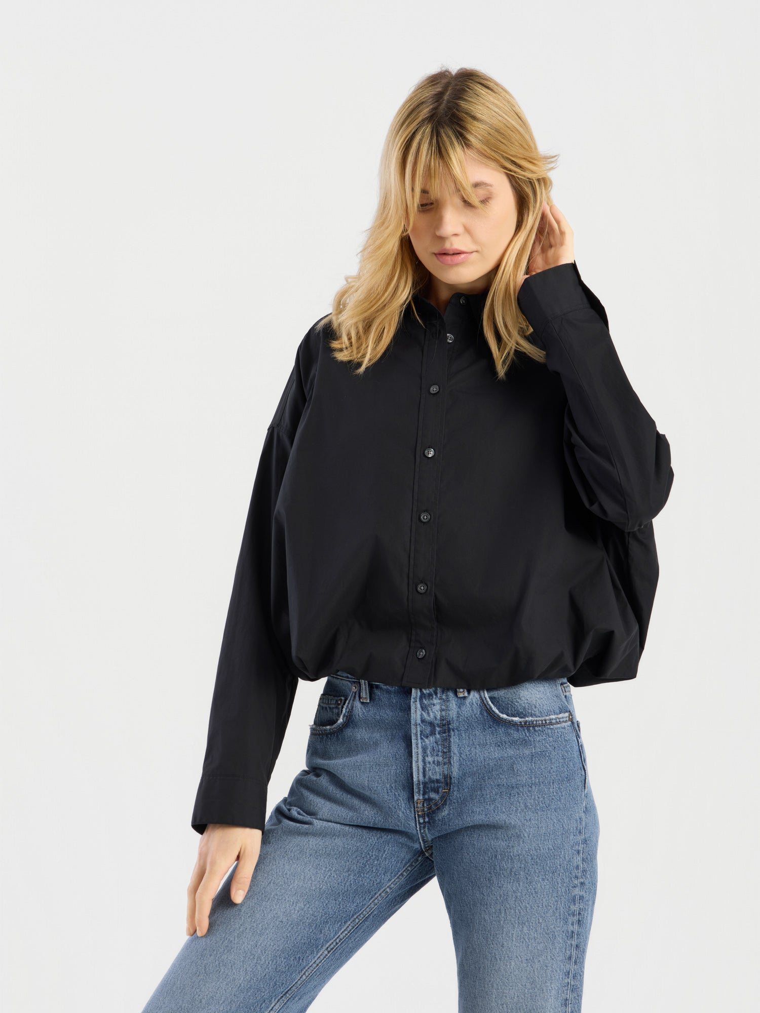 Eine Frau mit blonden Haaren trägt die „Ballon Blouse Black“, eine locker sitzende Bluse mit Knöpfen, dazu eine blaue Jeans. Sie steht vor einem schlichten weißen Hintergrund, berührt mit einer Hand ihr Haar und blickt nach unten.