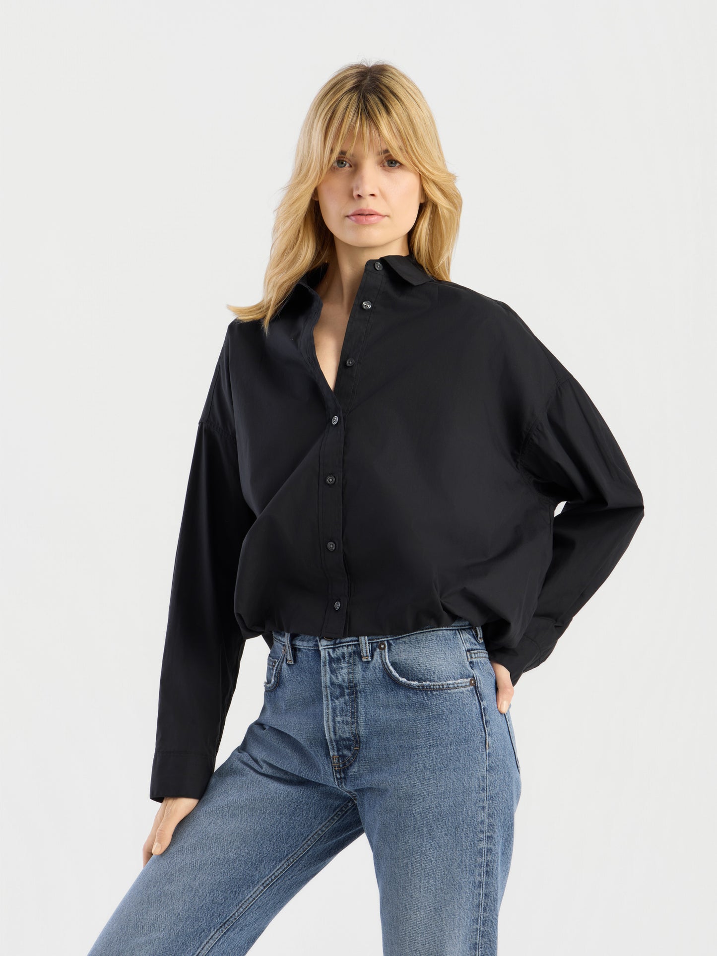 Eine Person mit blonden Haaren trägt die „Ballon Blouse Black“, ein locker sitzendes Hemd mit Knöpfen, dazu eine blaue Jeans, und steht mit einer Hand in der Hüfte vor einem schlichten, hellen Hintergrund.
