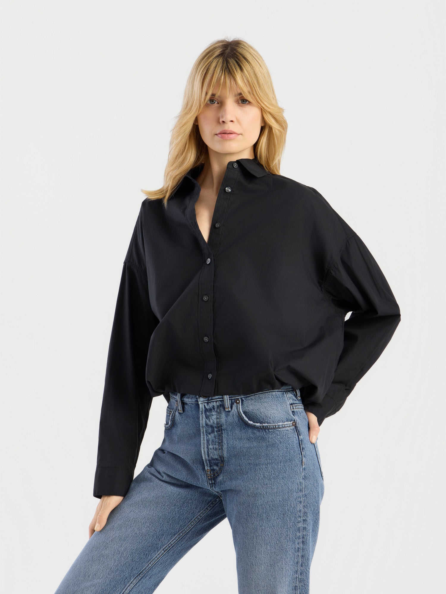 Eine Person mit blonden Haaren trägt die „Ballon Blouse Black“, ein locker sitzendes Hemd mit Knöpfen, dazu eine blaue Jeans, und steht mit einer Hand in der Hüfte vor einem schlichten, hellen Hintergrund.