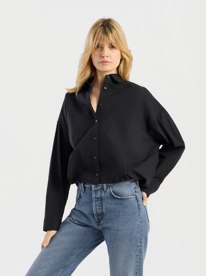 Eine Person mit blonden Haaren trägt die „Ballon Blouse Black“, ein locker sitzendes Hemd mit Knöpfen, dazu eine blaue Jeans, und steht mit einer Hand in der Hüfte vor einem schlichten, hellen Hintergrund.