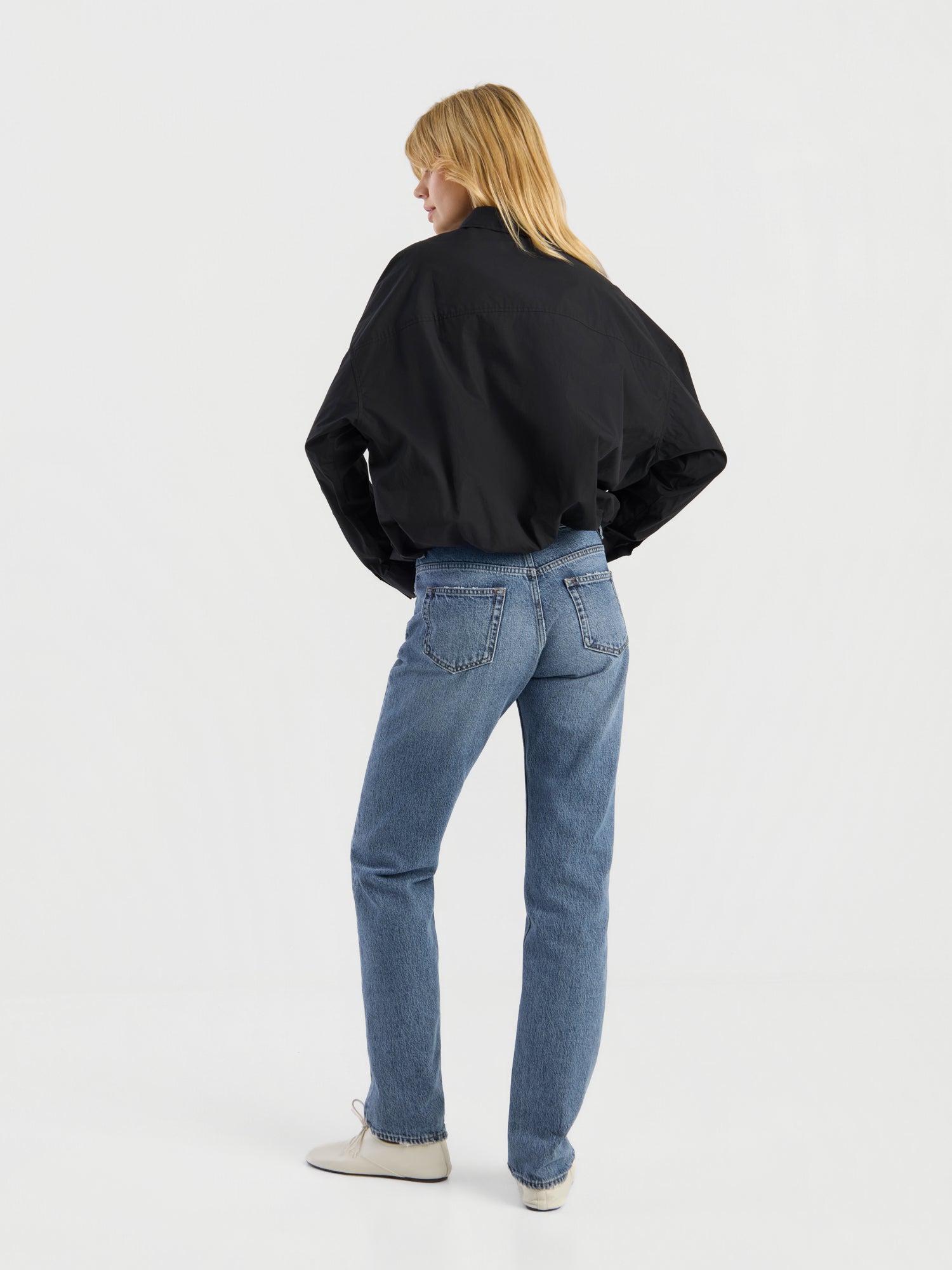 Eine Person mit blonden Haaren, die eine schwarze Jacke, eine „SIENNA Jeans Light Blue – Straight Leg Damenjeans“ und weiße Schuhe trägt, steht mit dem Rücken zur Kamera in einem minimalistisch gestalteten weißen Studio.