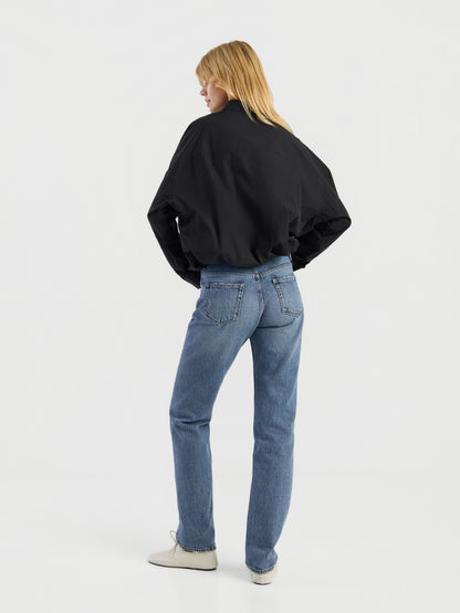 Eine Person mit blonden Haaren, die eine schwarze Jacke, eine „SIENNA Jeans Light Blue – Straight Leg Damenjeans“ und weiße Schuhe trägt, steht mit dem Rücken zur Kamera in einem minimalistisch gestalteten weißen Studio.