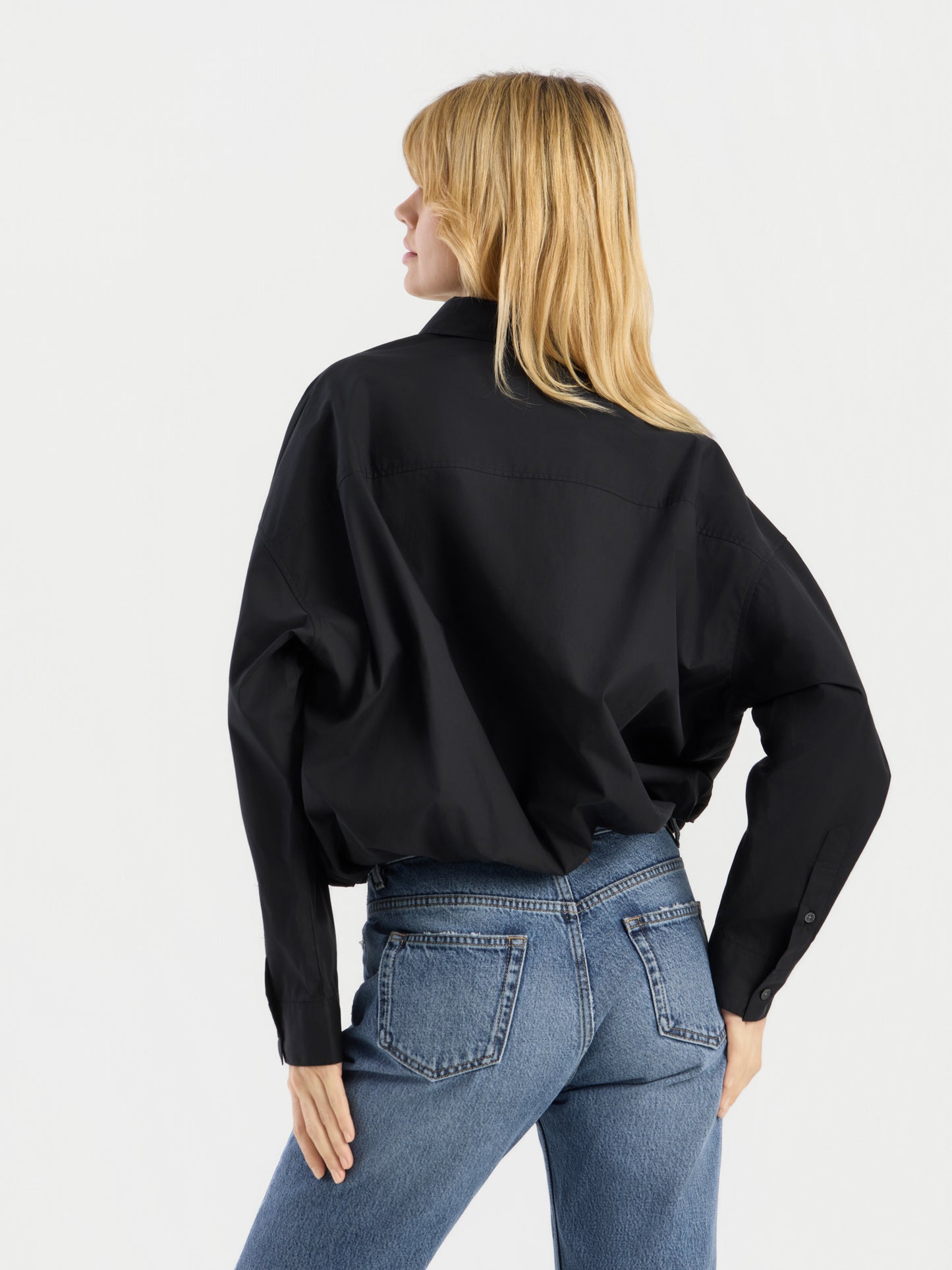 Eine Person mit langen blonden Haaren ist von hinten zu sehen; sie trägt die „Ballon Blouse Black“ und eine blaue Jeans vor einem schlichten weißen Hintergrund.