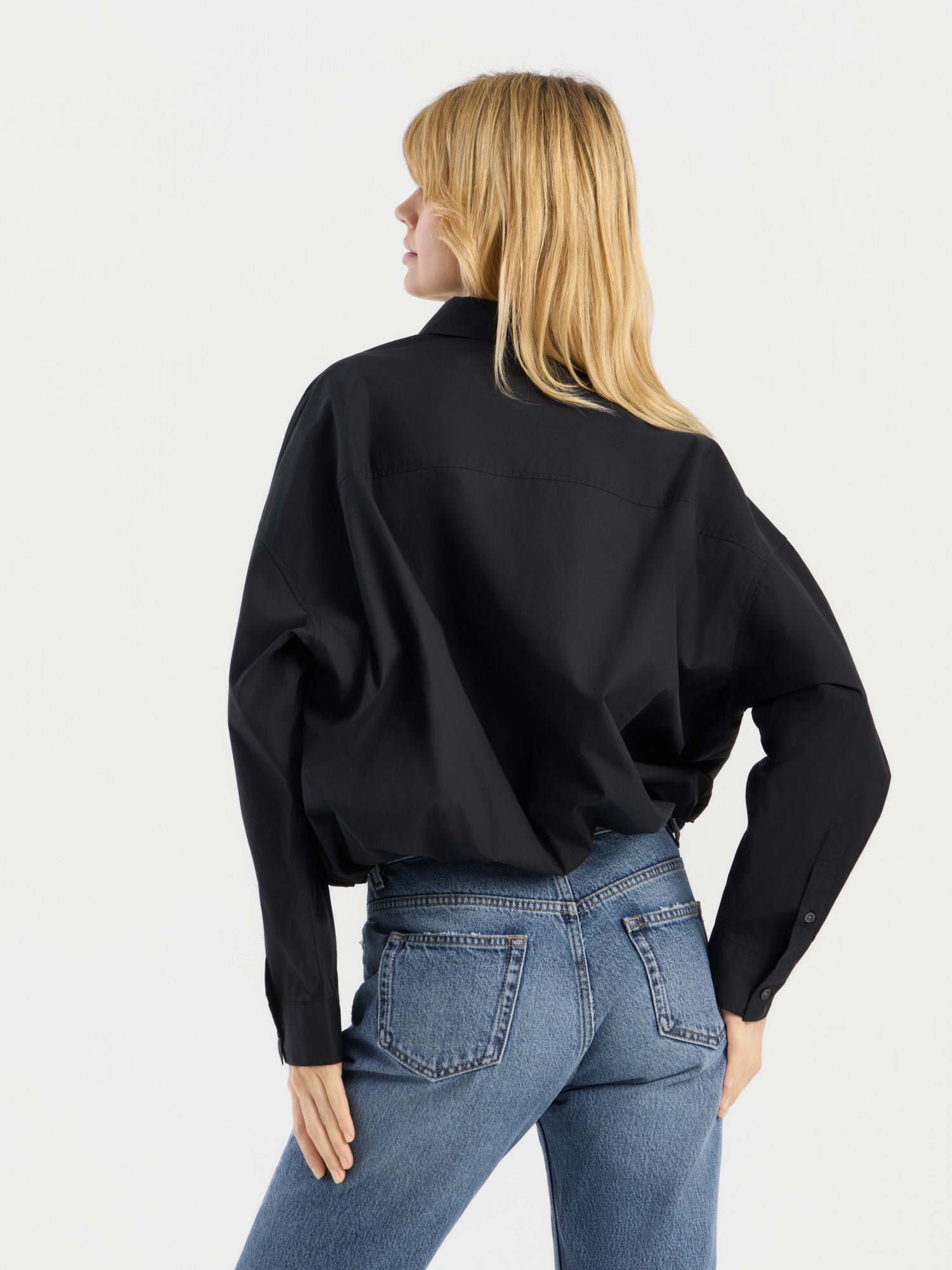 Eine Person mit langen blonden Haaren ist von hinten zu sehen; sie trägt die „Ballon Blouse Black“ und eine blaue Jeans vor einem schlichten weißen Hintergrund.