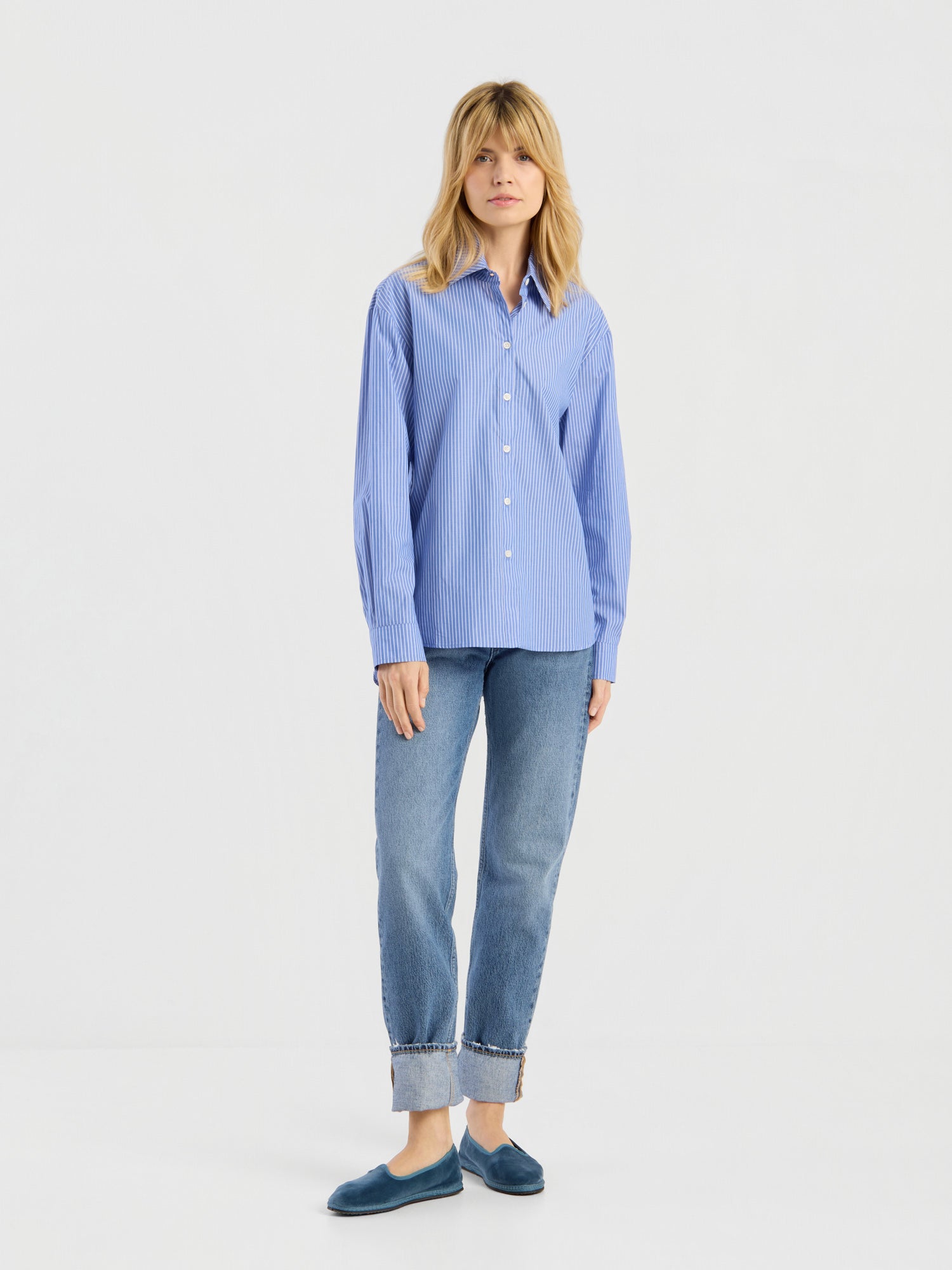 Vor einem schlichten weißen Hintergrund steht eine Person, die die Bluse „Emily“ – ein blau gestreiftes Hemd mit Knöpfen – sowie eine blaue Jeans mit Umschlag und blaue Slipper trägt.
