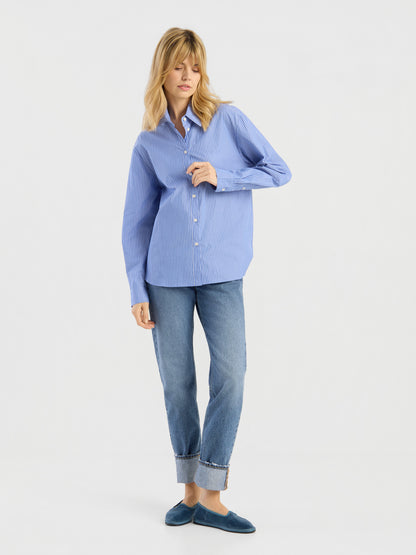 Eine Frau mit blonden Haaren trägt die Bluse „Emily“, ein blau gestreiftes Hemd mit Knöpfen, dazu eine blaue Jeans mit Umschlag und blaue Slipper. Sie steht vor einem schlichten weißen Hintergrund, blickt leicht zur Seite und hält eine Hand in der Nähe ihres Kragens.