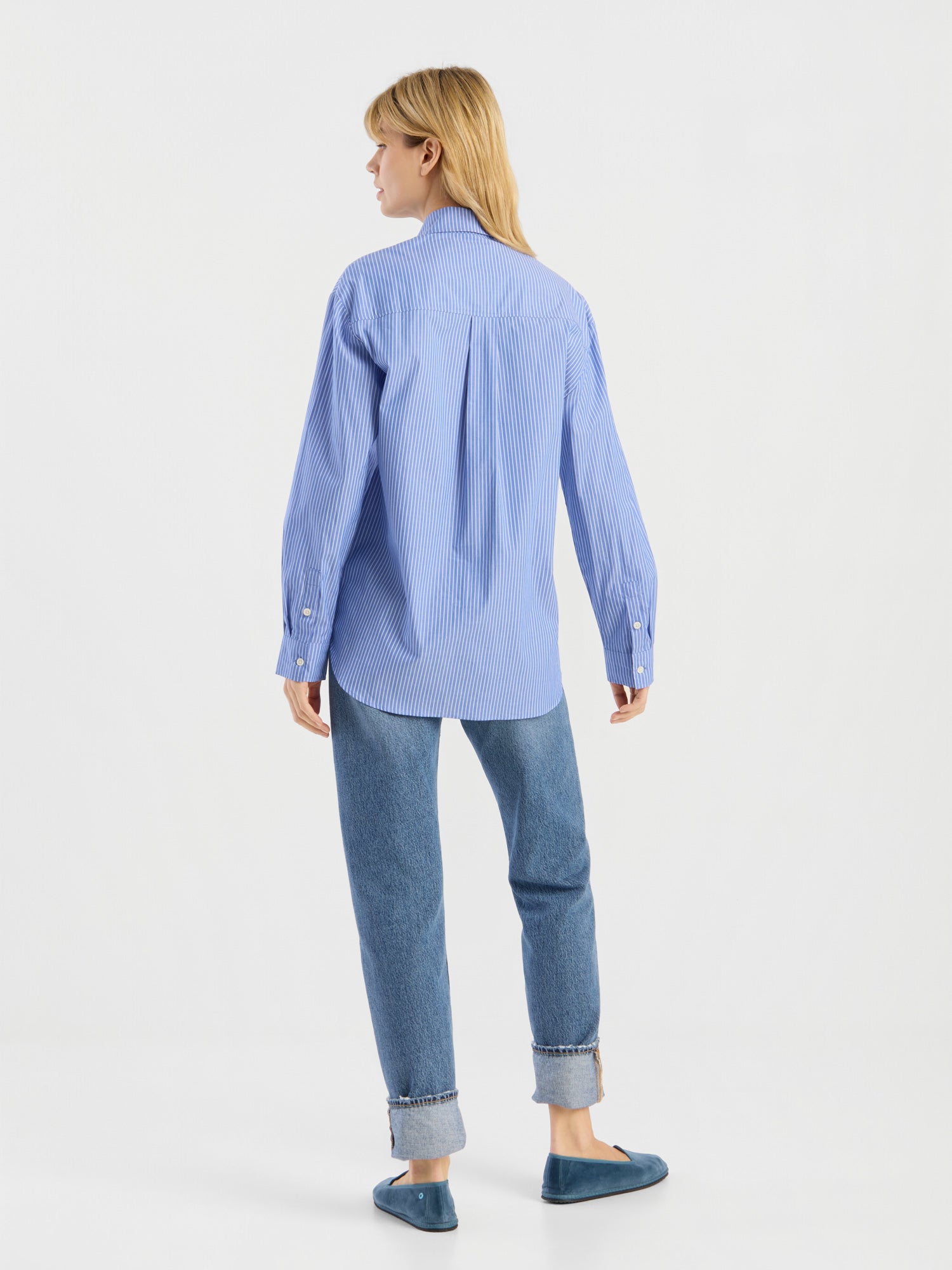 Vor einem schlichten weißen Hintergrund steht eine Person mit blonden Haaren mit dem Rücken zur Kamera. Sie trägt die „Bluse Emily“ – ein blau-weiß gestreiftes Hemd – sowie hochgekrempelte blaue Jeans und blaue Schuhe.