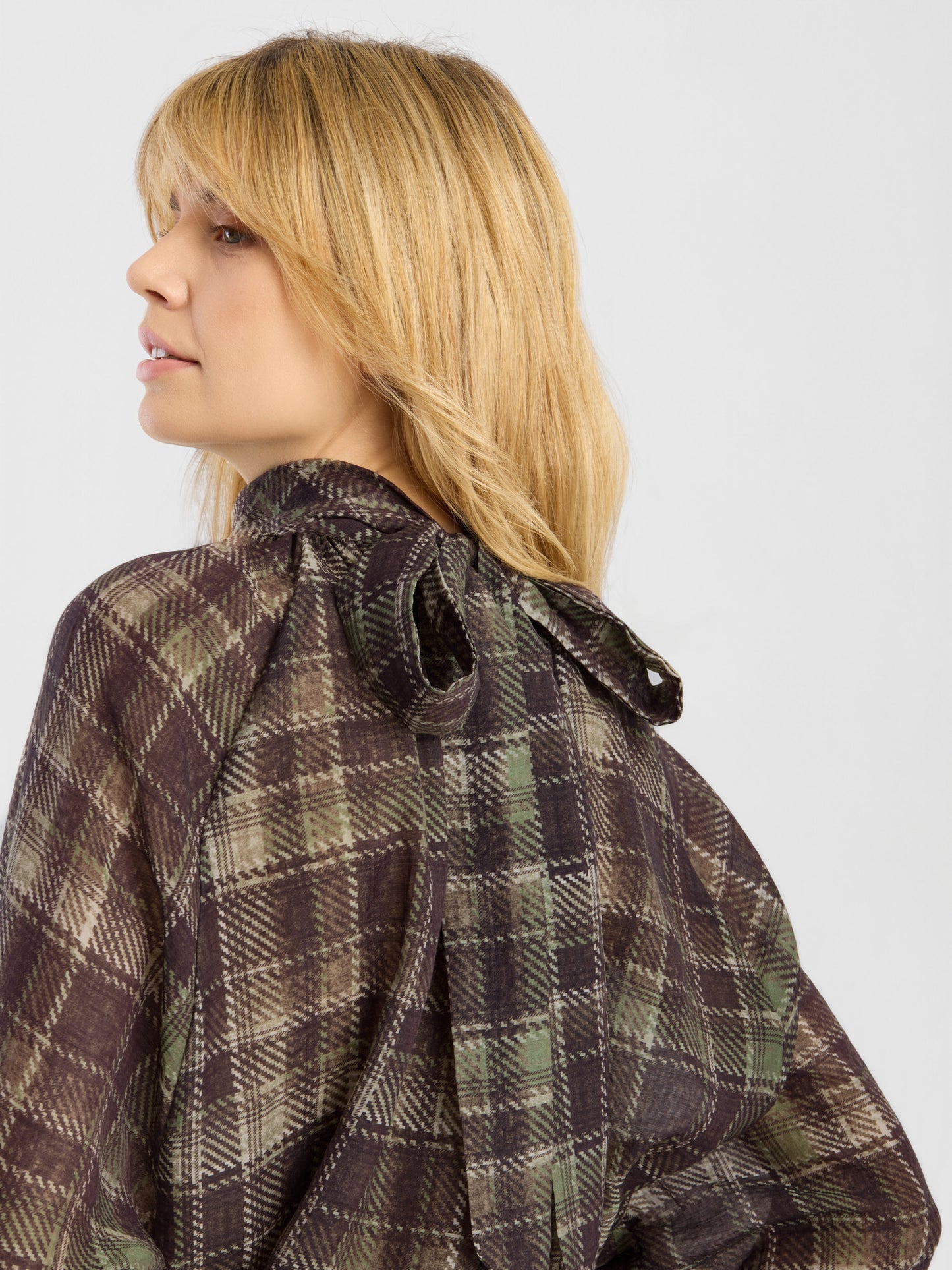 Eine Person mit glattem, schulterlangem blondem Haar präsentiert das Modell „Bluse Bow Check“ – ein braun-grün kariertes Oberteil mit einer Schleife im Nacken – und blickt dabei vor einem einfarbigen Hintergrund über die Schulter.