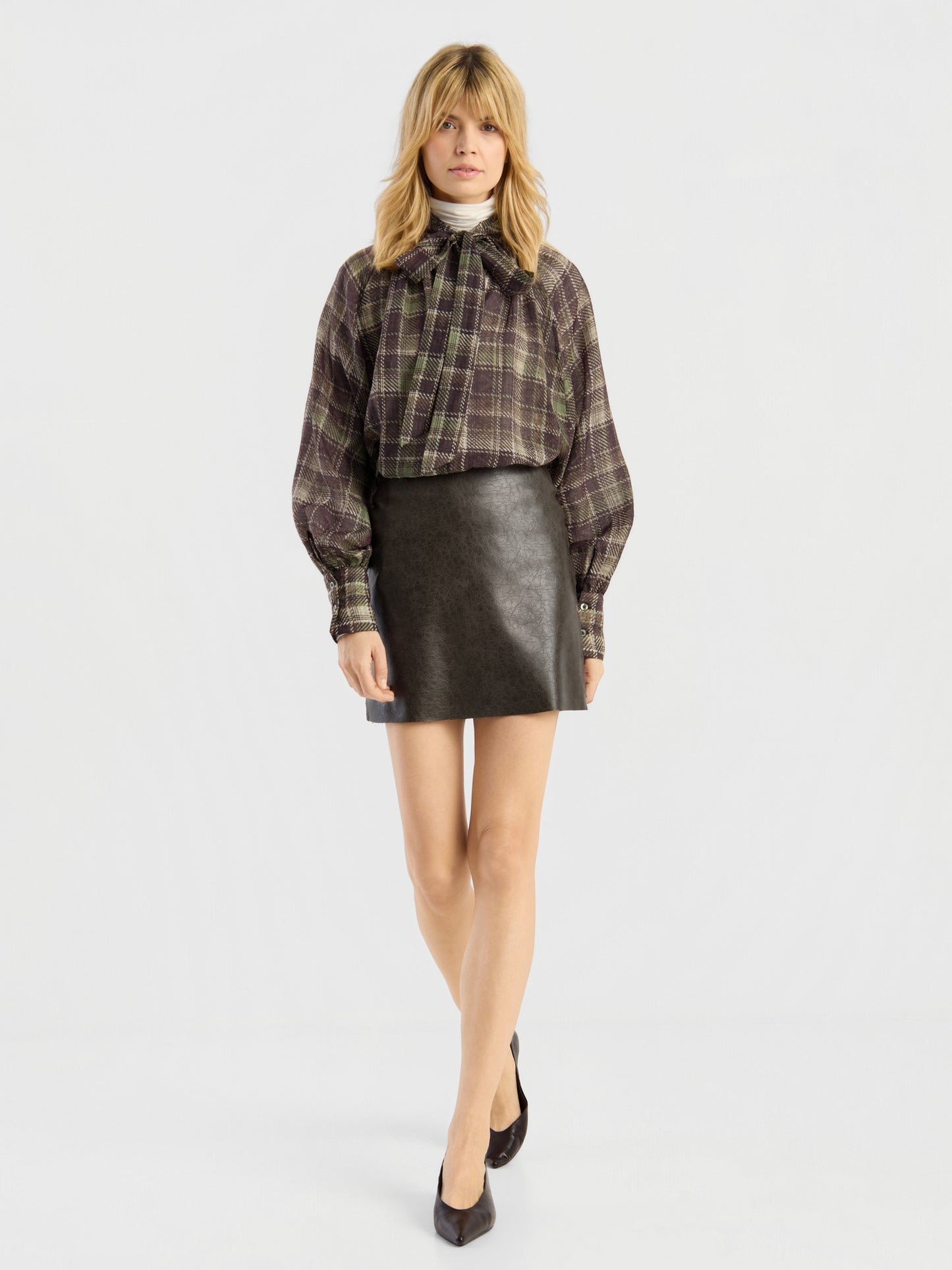 Eine Frau mit blonden Haaren steht vor einem schlichten weißen Hintergrund, den Blick nach vorne gerichtet, und trägt das „Bluse Bow Check“-Hemd, einen schwarzen Lederrock und schwarze Pumps.