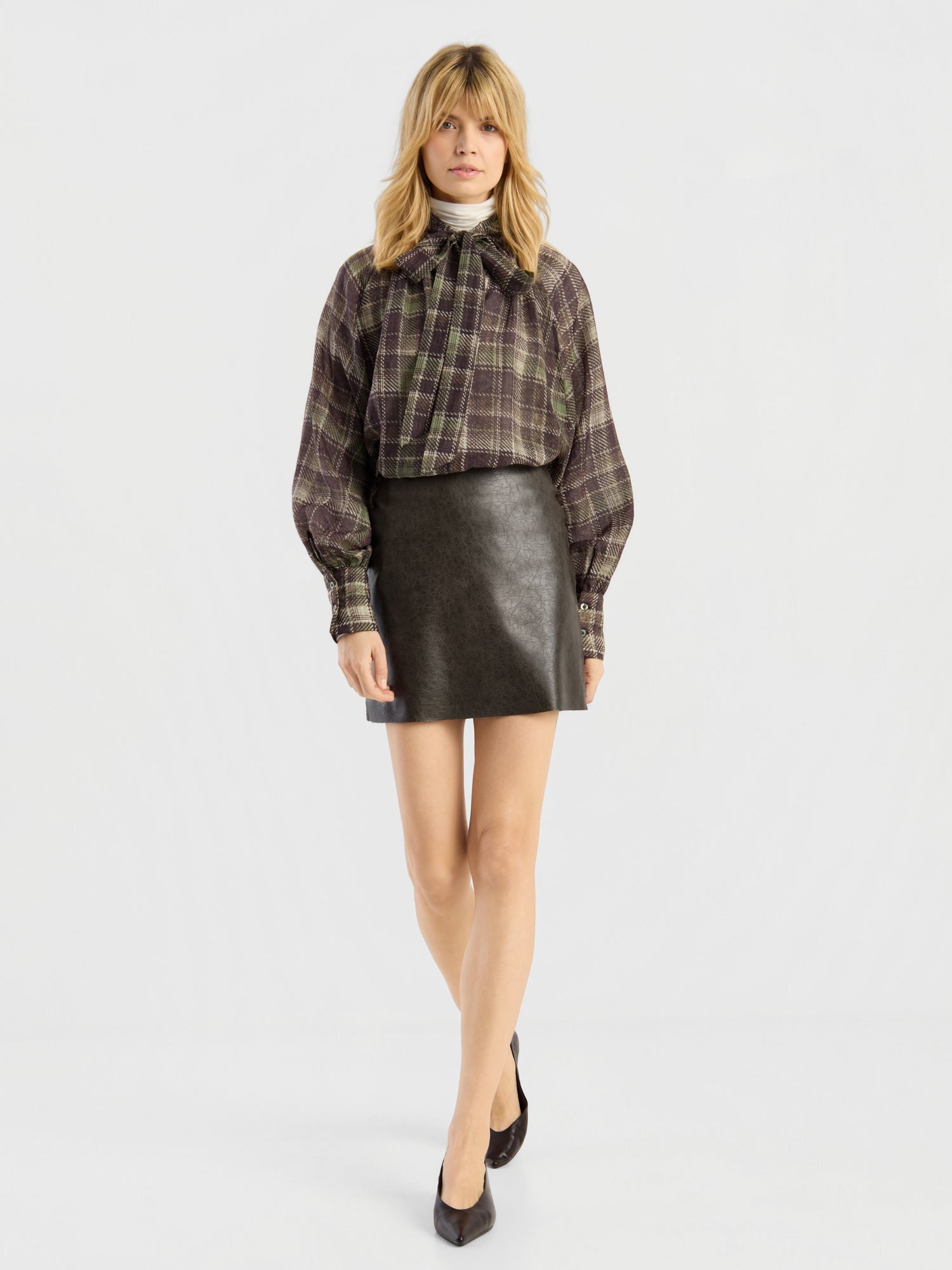 Eine Frau, die ein kariertes Hemd, den „Mini Skirt 70’s“ aus schwarzem Leder und schwarze Pumps trägt, steht vor einem schlichten weißen Hintergrund.
