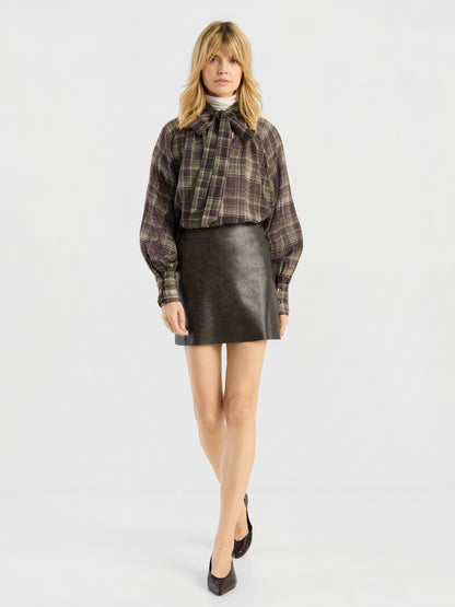 Eine Frau, die ein kariertes Hemd, den „Mini Skirt 70’s“ aus schwarzem Leder und schwarze Pumps trägt, steht vor einem schlichten weißen Hintergrund.