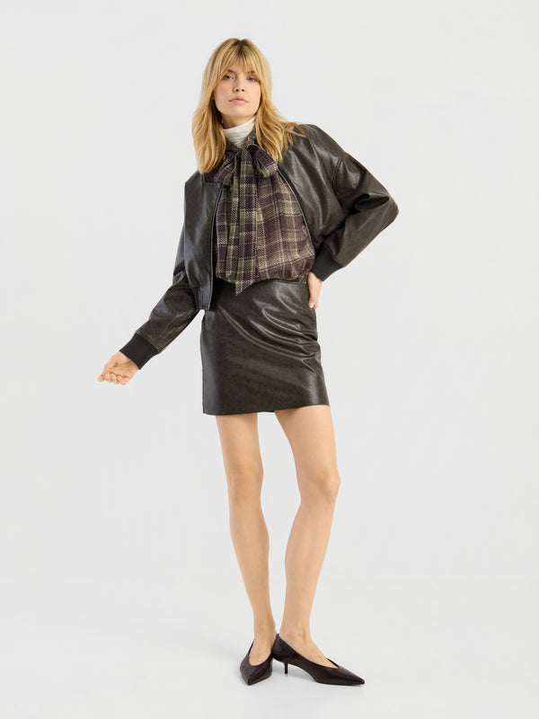 Eine Frau präsentiert den „Vegan-Leder-Look“ – eine schwarze Kunstlederjacke und einen dazu passenden Minirock –, kombiniert mit einer auffälligen karierten Bluse mit Schleife und schwarzen Pumps, vor einem schlichten, hellen Hintergrund.