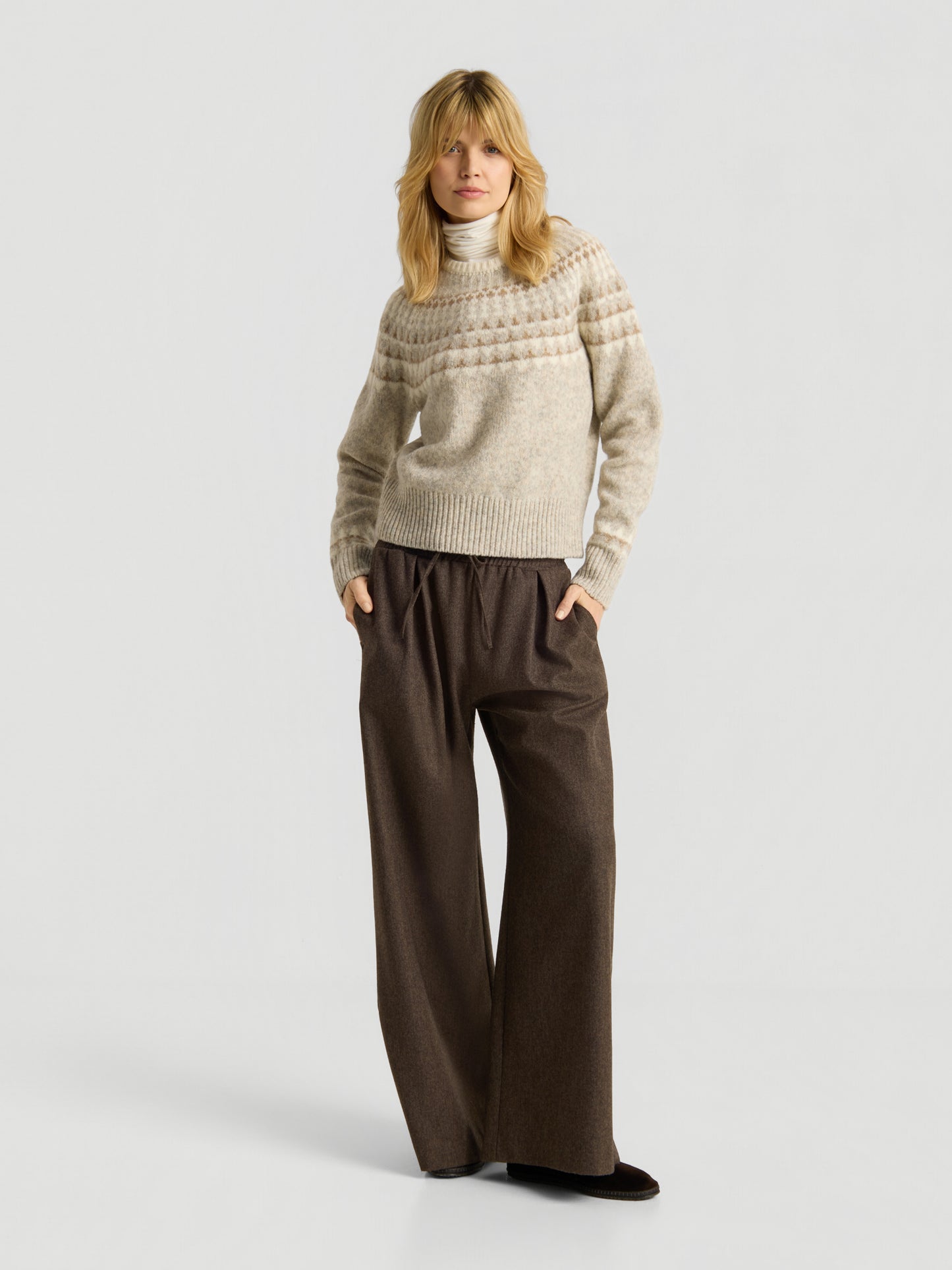 Eine Person trägt den „Norweger Greige Vol. 3“ und steht mit den Händen in den Taschen vor einem schlichten, hellen Hintergrund. Sie trägt einen strukturierten, beigen Strickpullover und eine lockere, weit geschnittene braune Hose und blickt dabei leicht zur Seite.