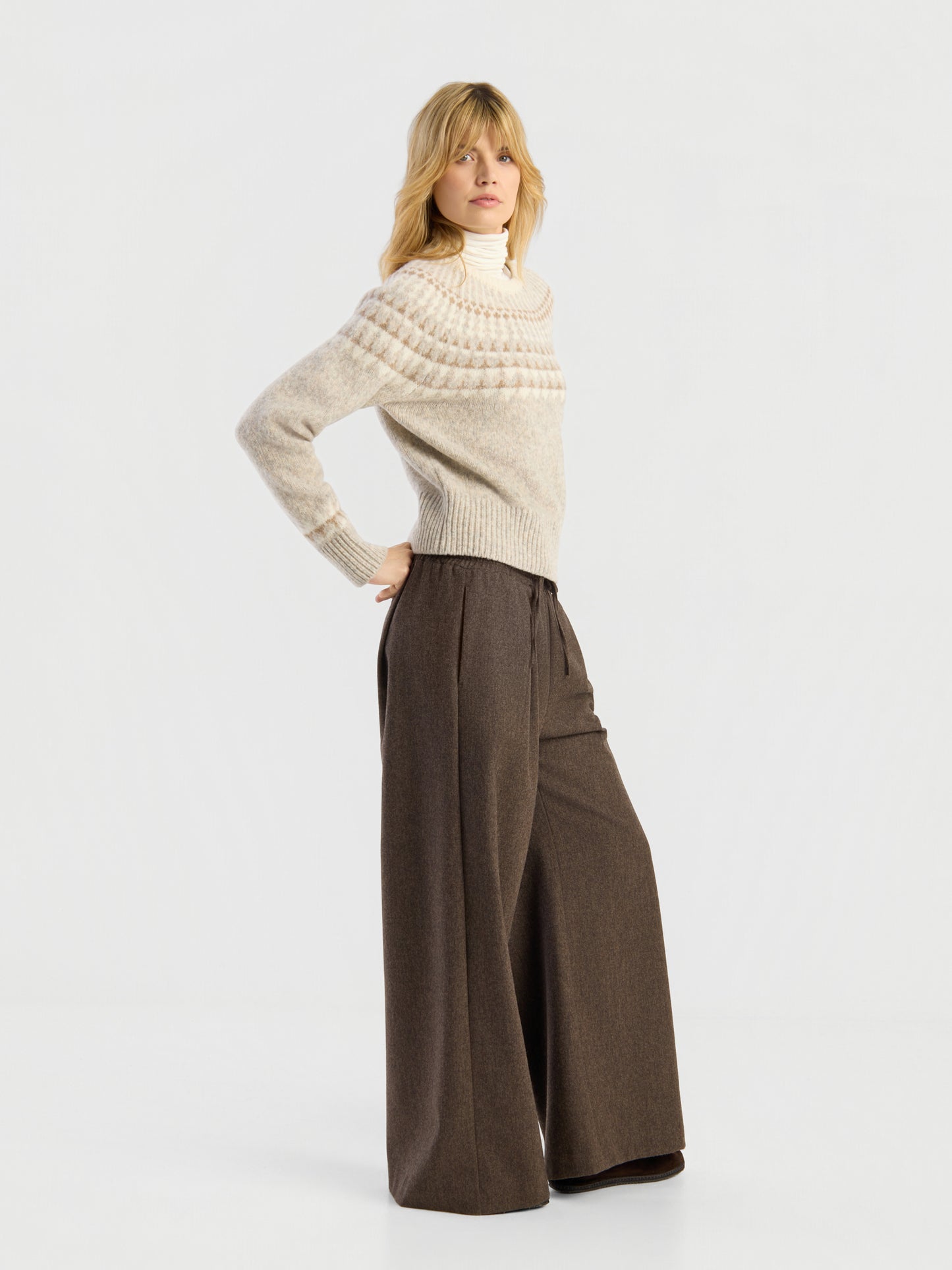 Eine Person steht vor einem schlichten weißen Hintergrund und posiert mit einer Hand in der Hüfte. Sie trägt den Pullover „Norweger Greige Vol. 3“ und eine braune Hose mit hoher Taille und weitem Bein.