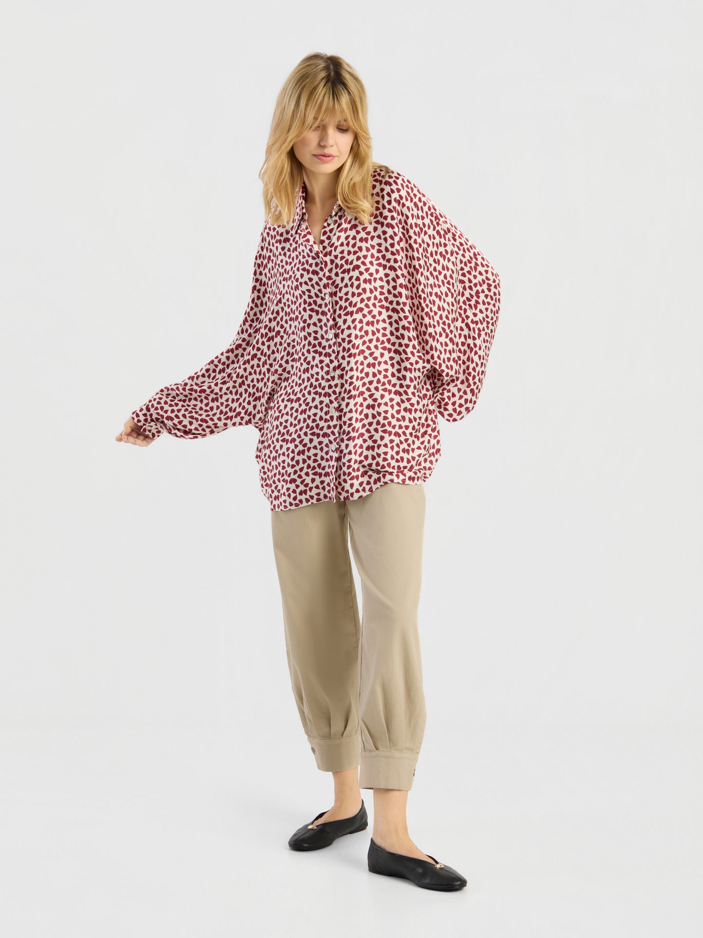 Eine Frau mit blonden Haaren steht in einer entspannten Haltung vor einem weißen Hintergrund. Sie trägt ein rot-weiß gemustertes Langarmshirt von Antonia Valentine, eine beige Hose mit Umschlag und schwarze Ballerinas.