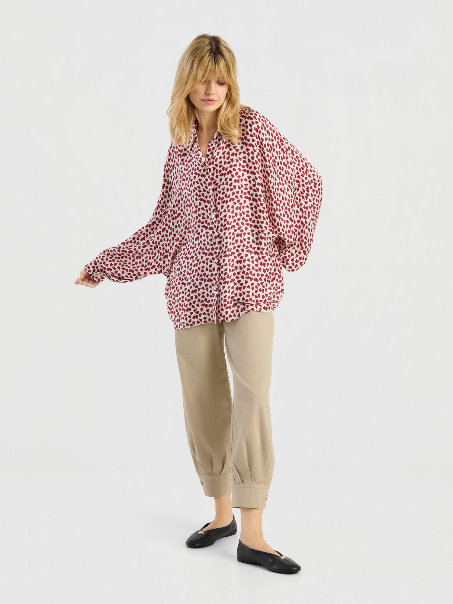 Eine Frau mit blonden Haaren steht in einer entspannten Haltung vor einem weißen Hintergrund. Sie trägt ein rot-weiß gemustertes Langarmshirt von Antonia Valentine, eine beige Hose mit Umschlag und schwarze Ballerinas.