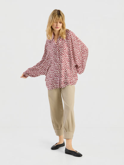 Eine Frau mit blonden Haaren steht in einer entspannten Haltung vor einem weißen Hintergrund. Sie trägt ein rot-weiß gemustertes Langarmshirt von Antonia Valentine, eine beige Hose mit Umschlag und schwarze Ballerinas.