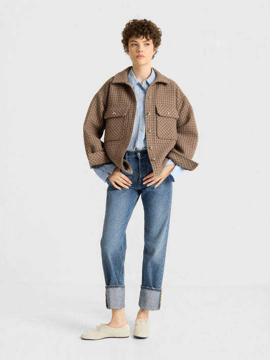 Eine Person mit kurzen Locken trägt die braune Karojacke mit der Aufschrift „Check, Please!“ über einem hellblauen Hemd, dazu blaue Jeans mit Umschlag und weiße Schuhe, und steht vor einem schlichten hellen Hintergrund.